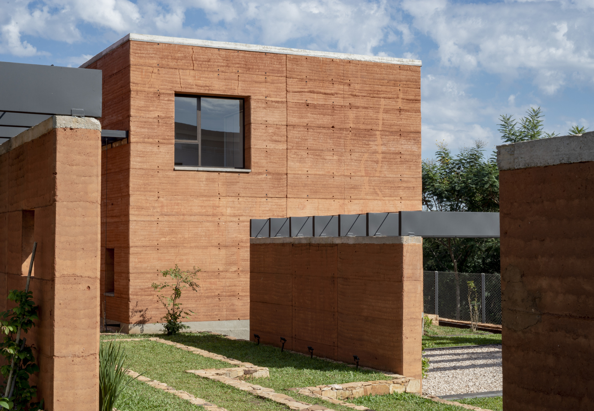 Two-storey rammed earth volume with metal bridge connection under a cloudy afternoon sky