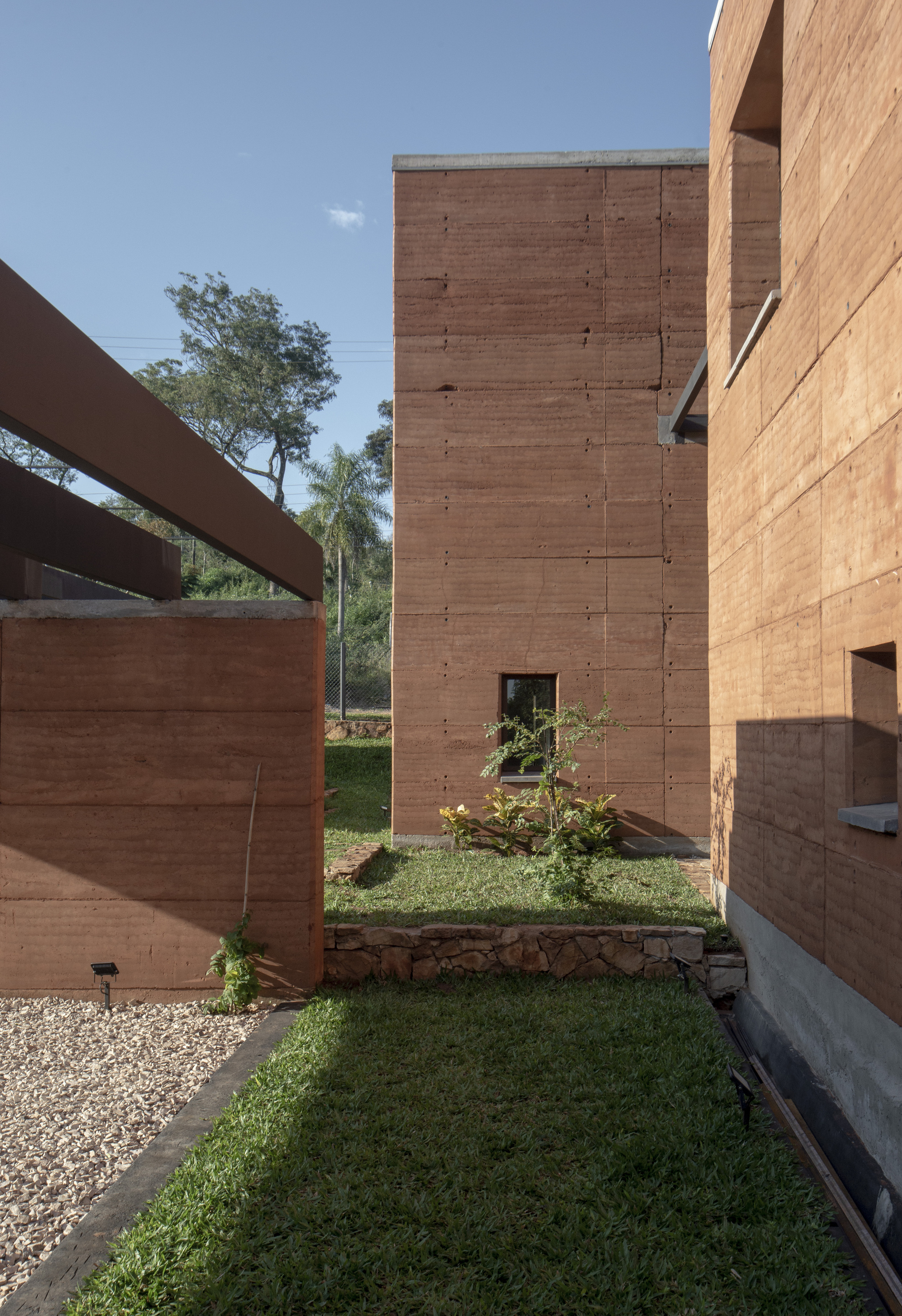 Narrow garden passage between rammed earth volumes with grass strips and young planted trees