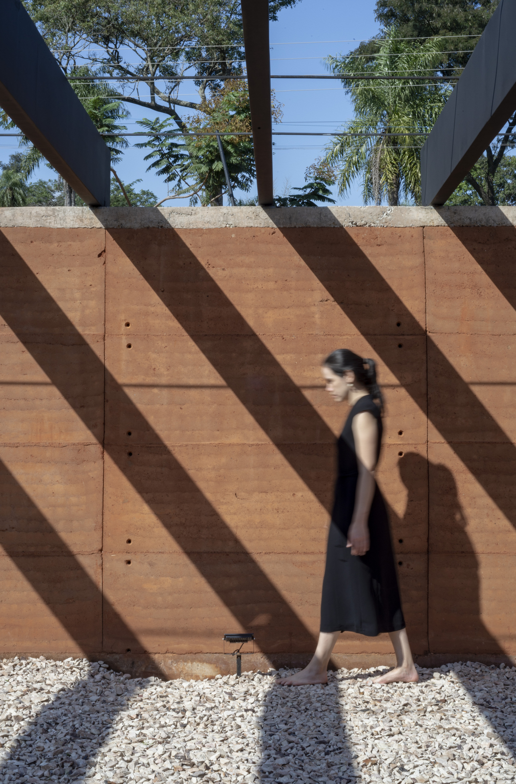 Figure walking past rammed earth wall with diagonal shadows cast by overhead steel beams on gravel