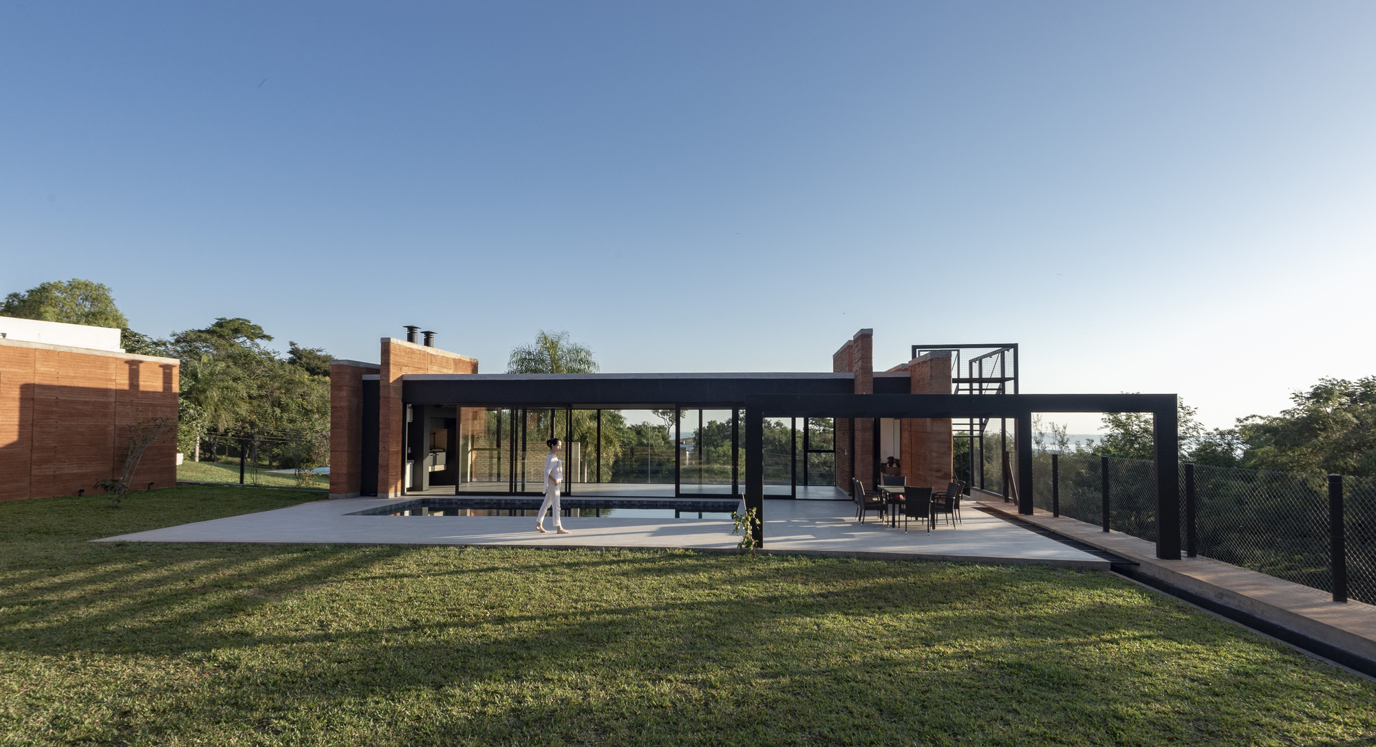 Glass pavilion with steel frame connecting rammed earth volumes across a sloped lawn at dusk