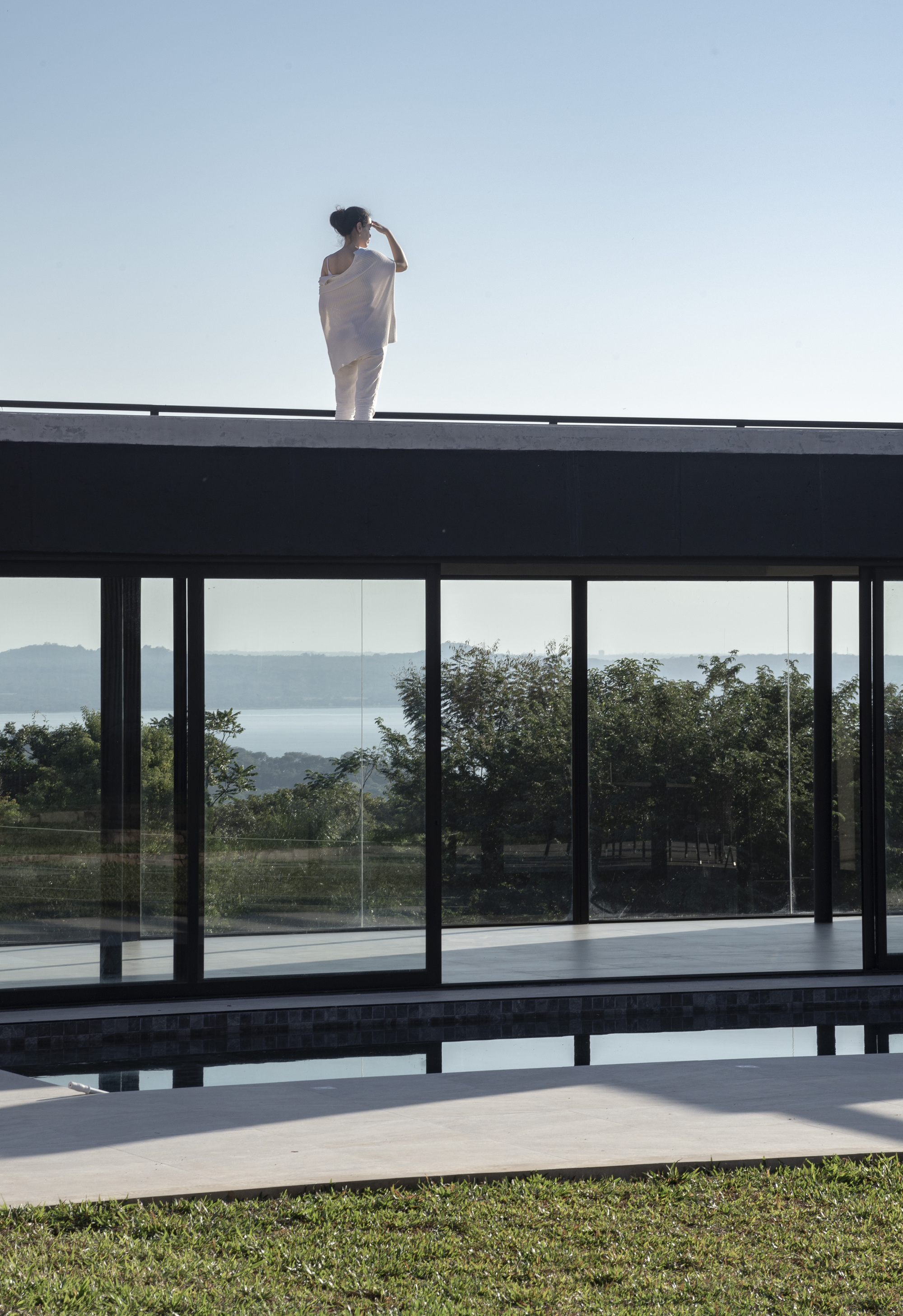 Figure standing on the flat roof above the glass-walled pool pavilion overlooking distant hills
