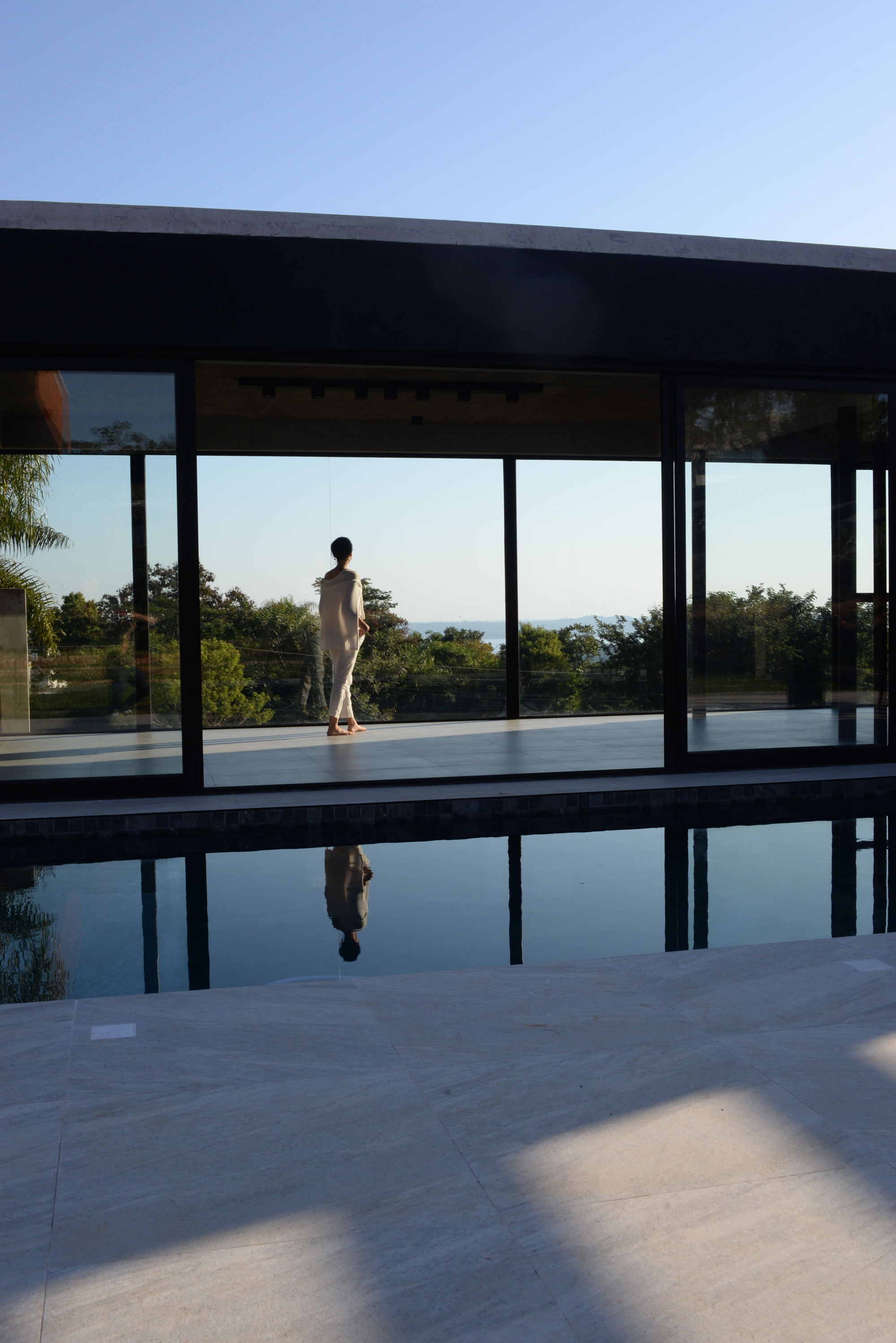 Pool terrace with figure reflected in still water beside the steel-framed glass pavilion at sunset