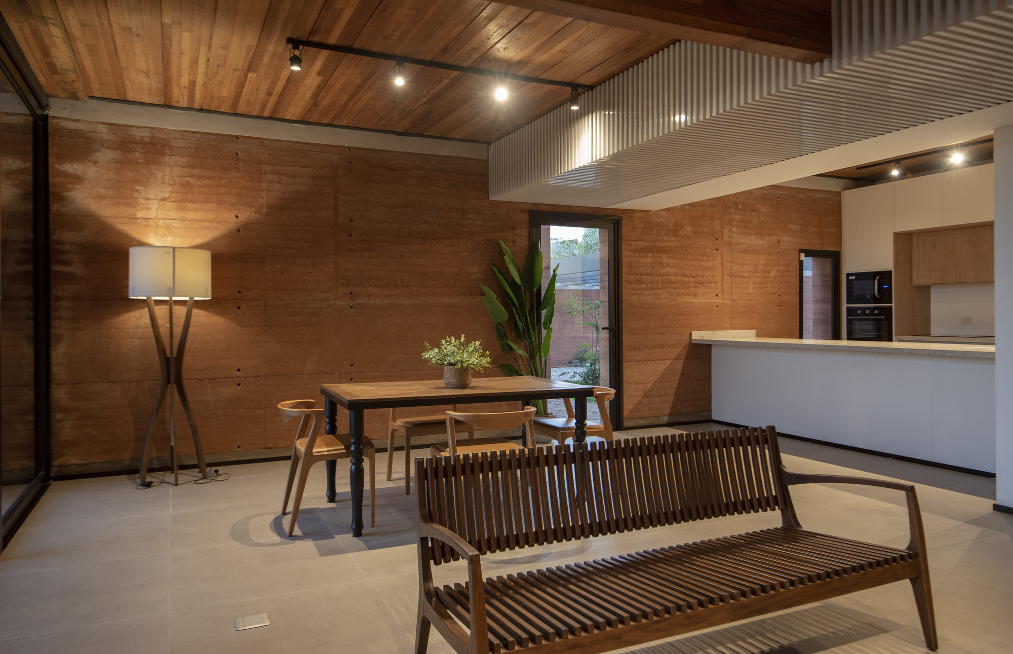 Open-plan living and dining space with rammed earth walls, timber ceiling and corrugated metal air duct