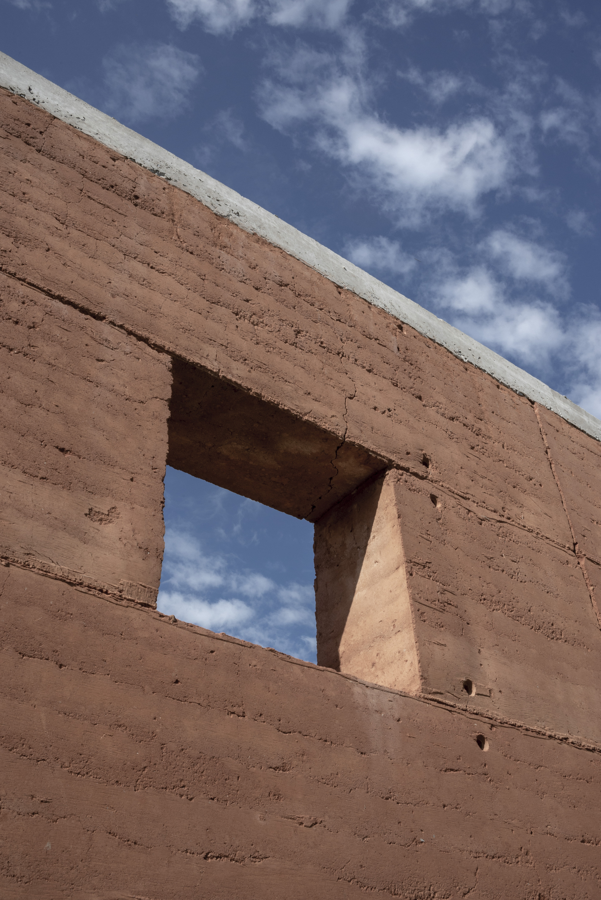 Rammed earth wall with rectangular window opening framing blue sky and white clouds