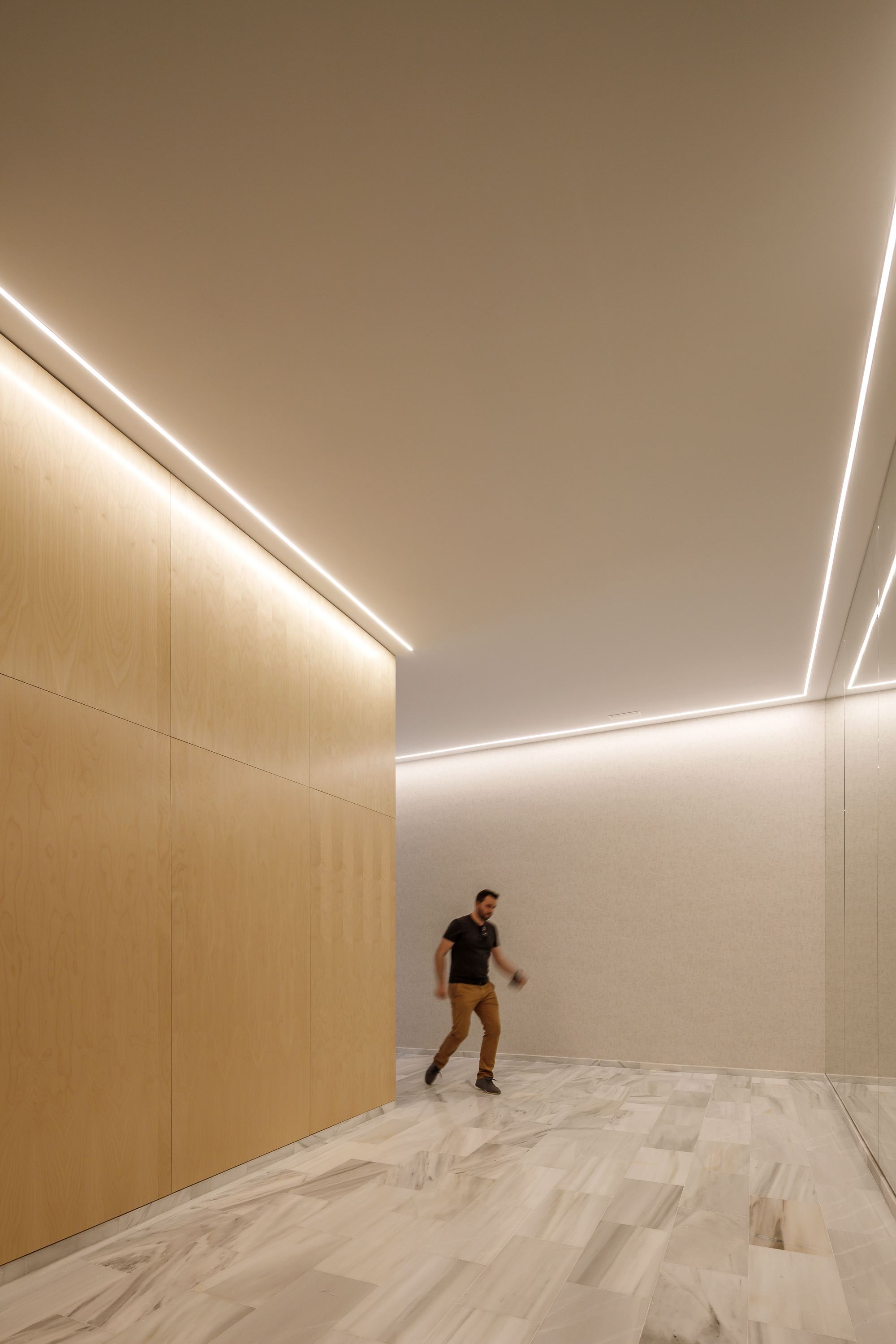 Interior corridor with concealed cove lighting along timber and white plaster walls, one person walking