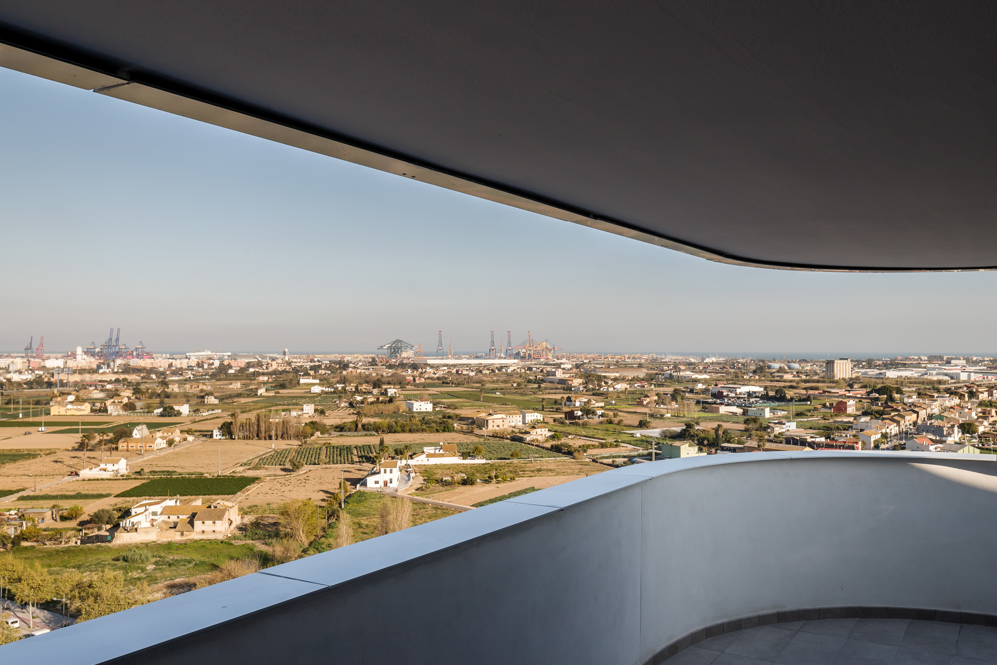 Curved balcony edge beneath dark overhang overlooking rural landscape with industrial skyline in the distance