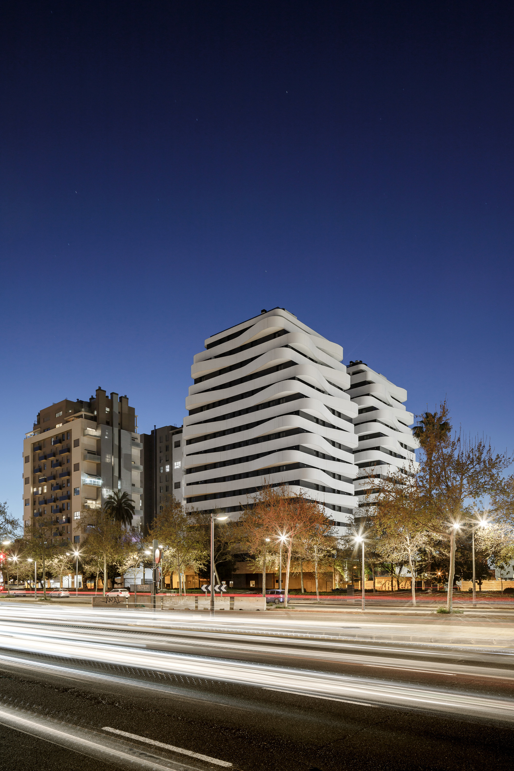 Street view of the stacked balcony facade at twilight with traffic light trails and illuminated trees