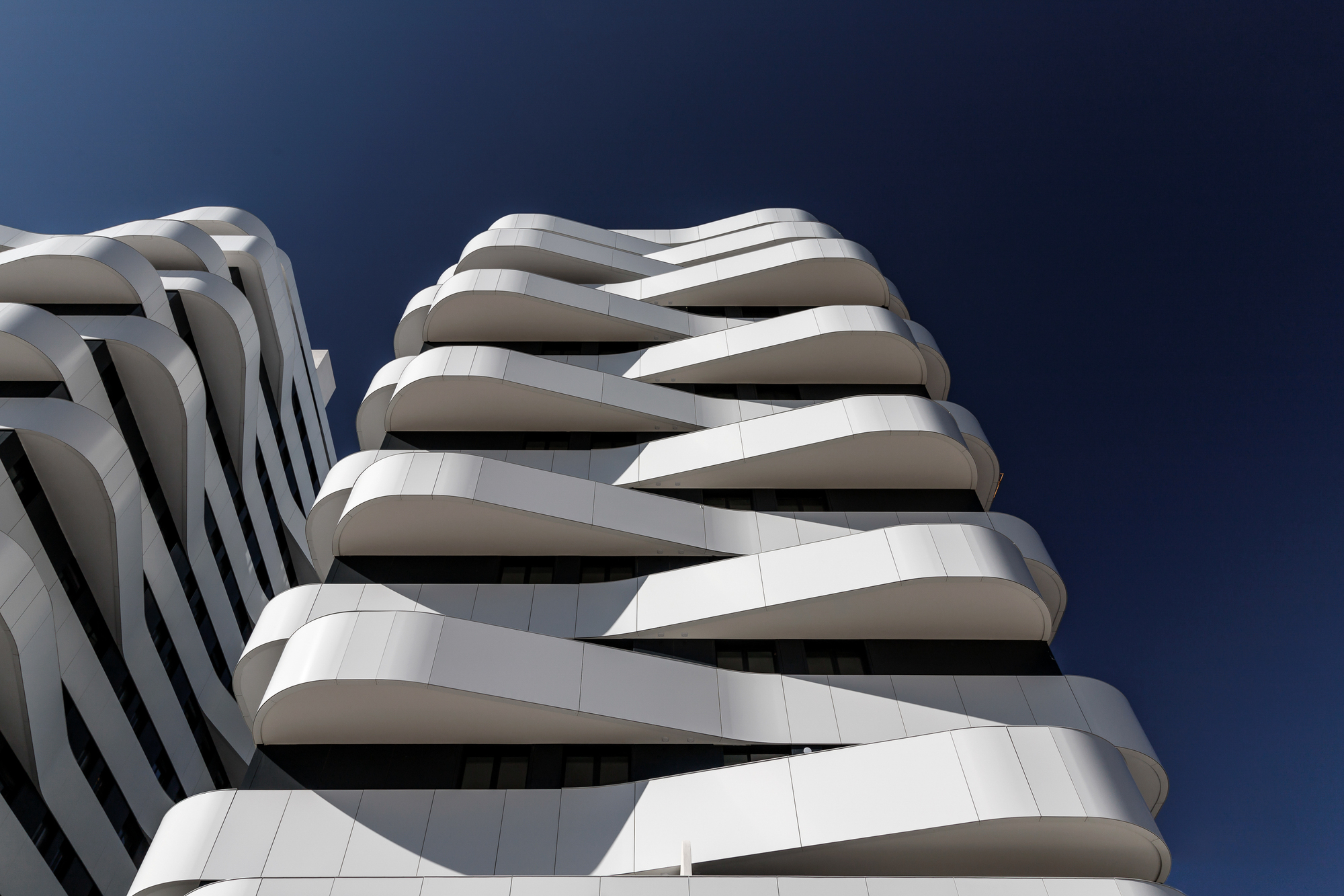 Upward view of the curving white balcony slabs wrapping the tower facade under bright sunlight