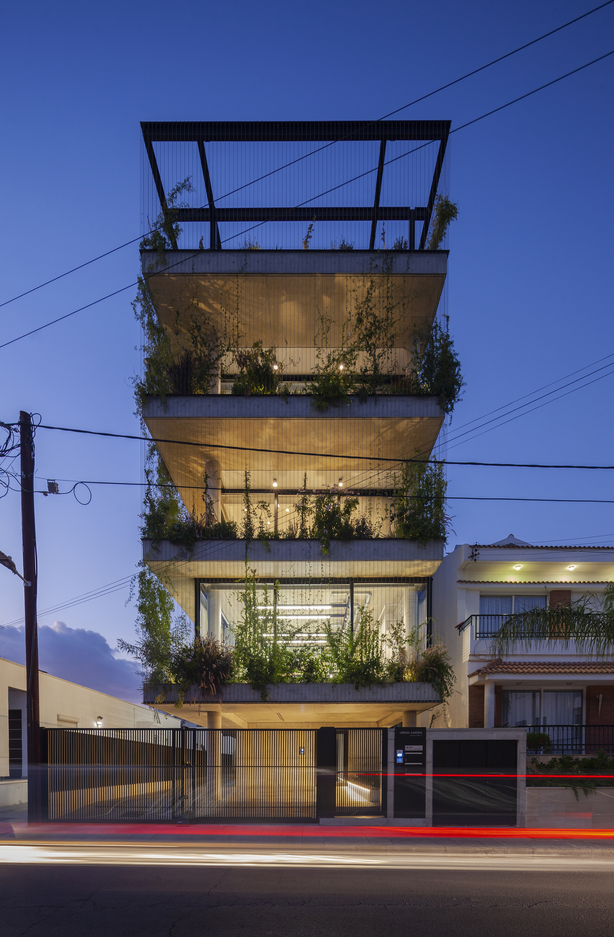 Street view of a four-story concrete facade with planted balconies at dusk with car light trails