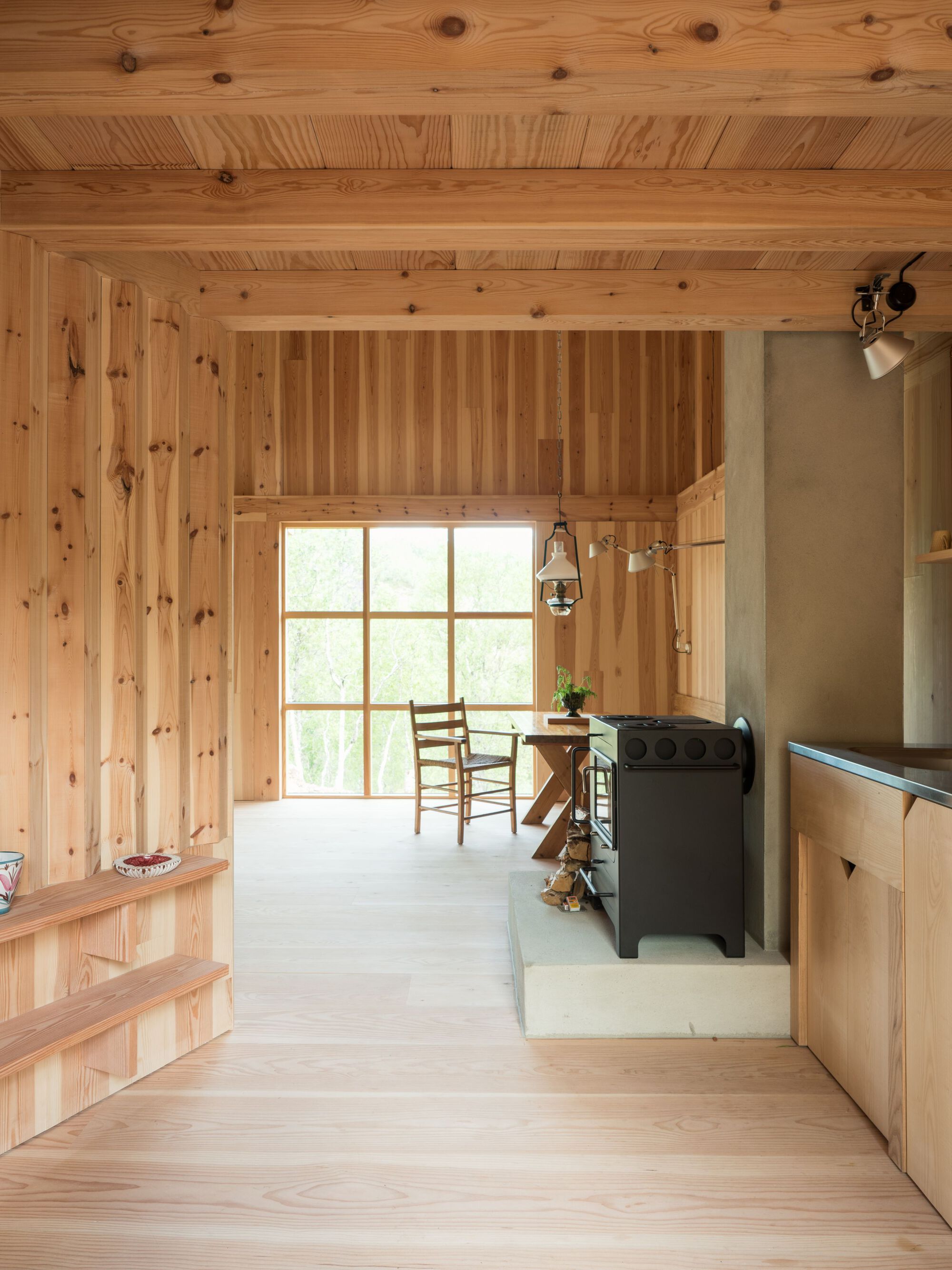 Interior with wood-lined walls and ceiling, large gridded window and wood-burning stove in the corner