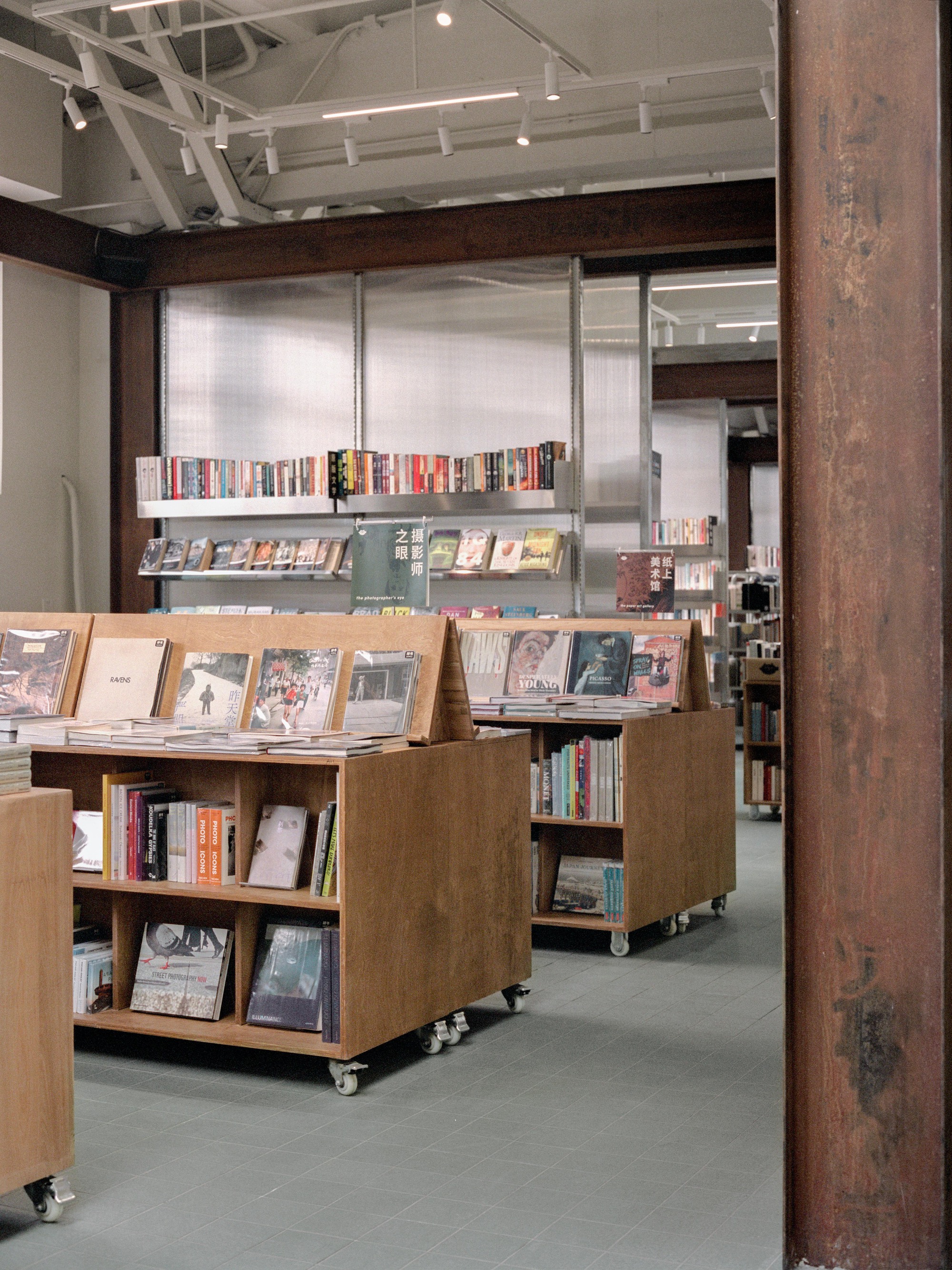 Wheeled plywood display units holding books in front of translucent wall panels and metal shelves beyond timber column