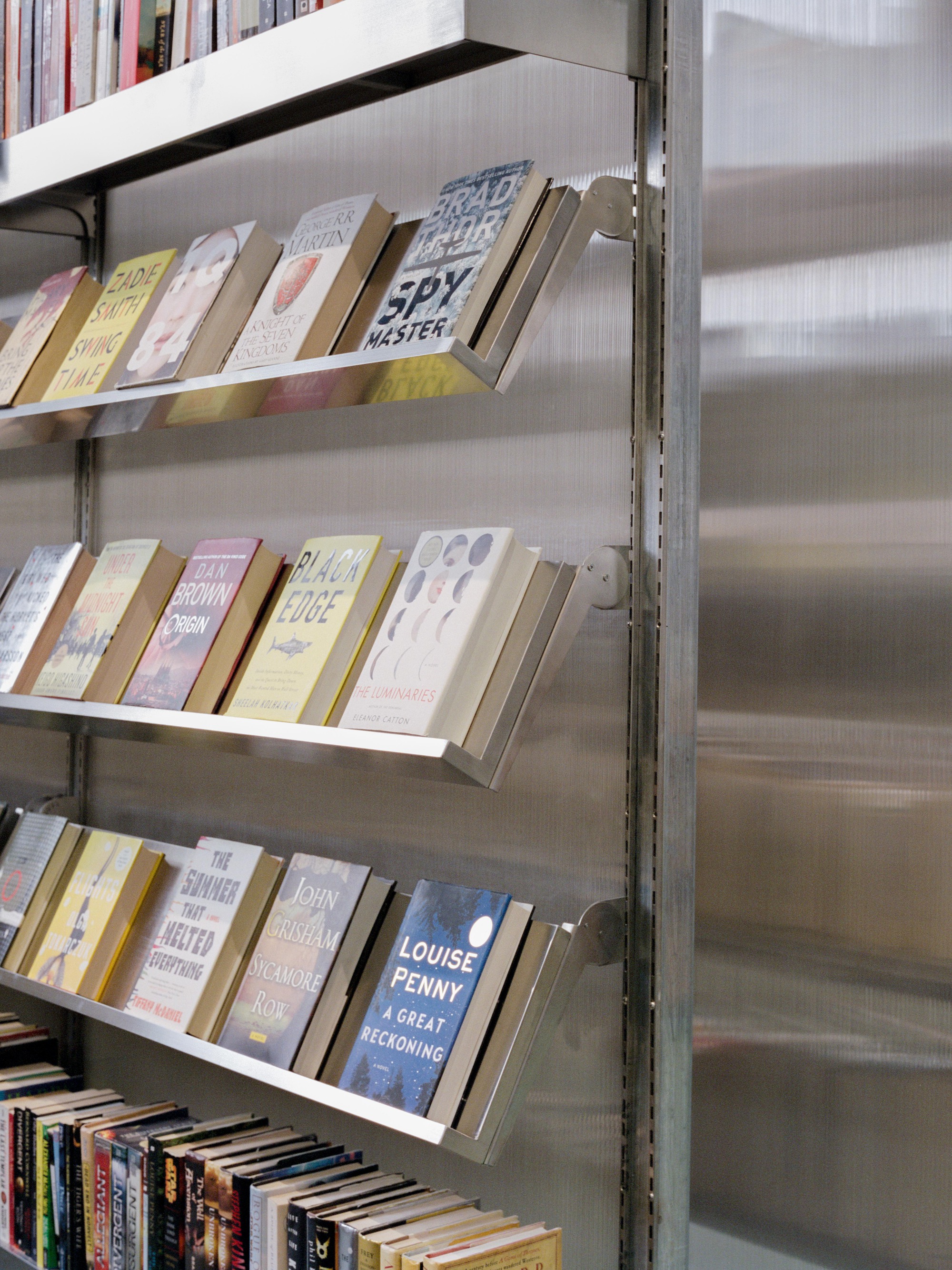 Metal bookshelves with angled display ledges holding books against a corrugated polycarbonate wall