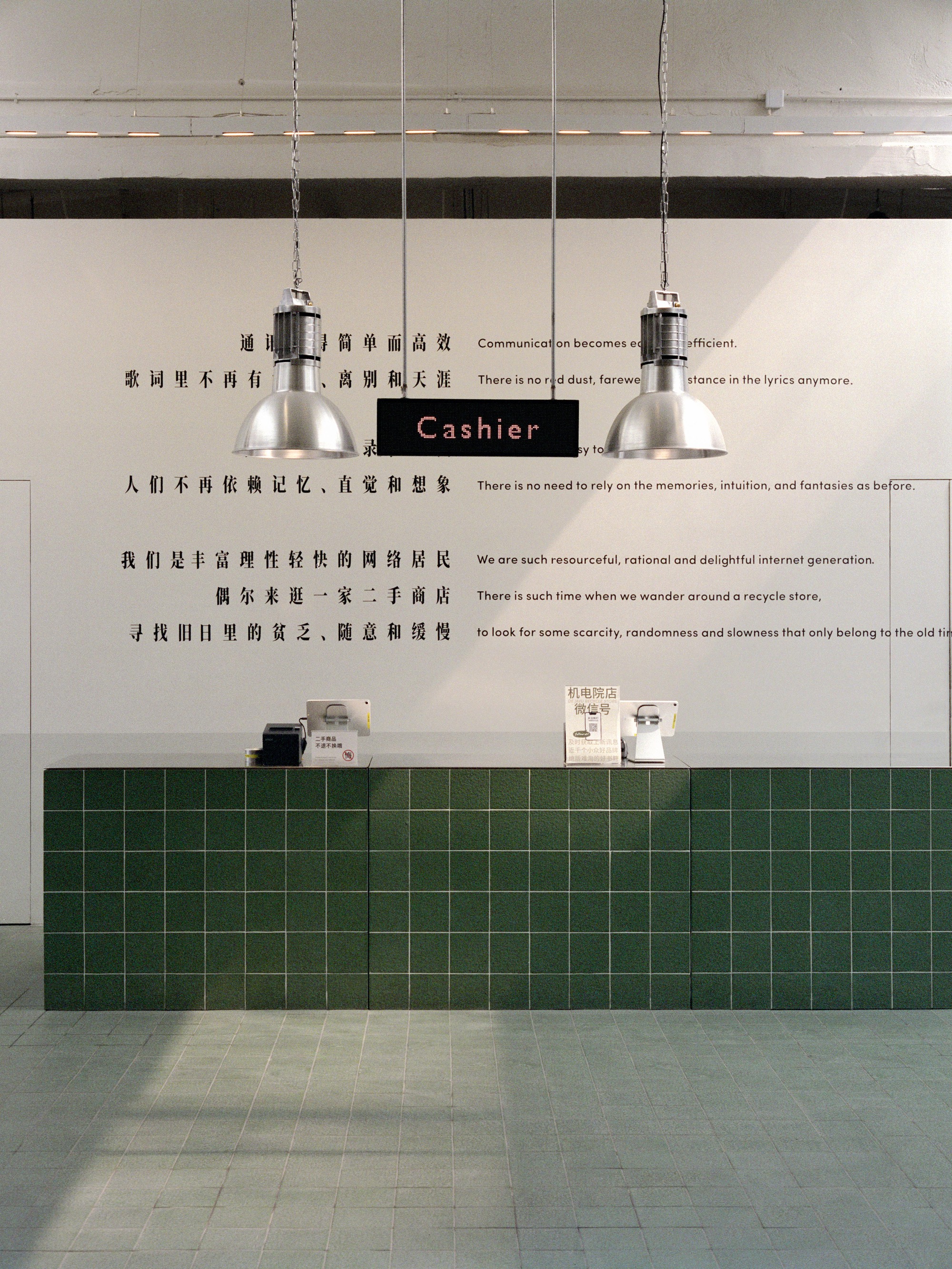 Cashier counter clad in dark green square tiles beneath two chrome pendant lights and wall text