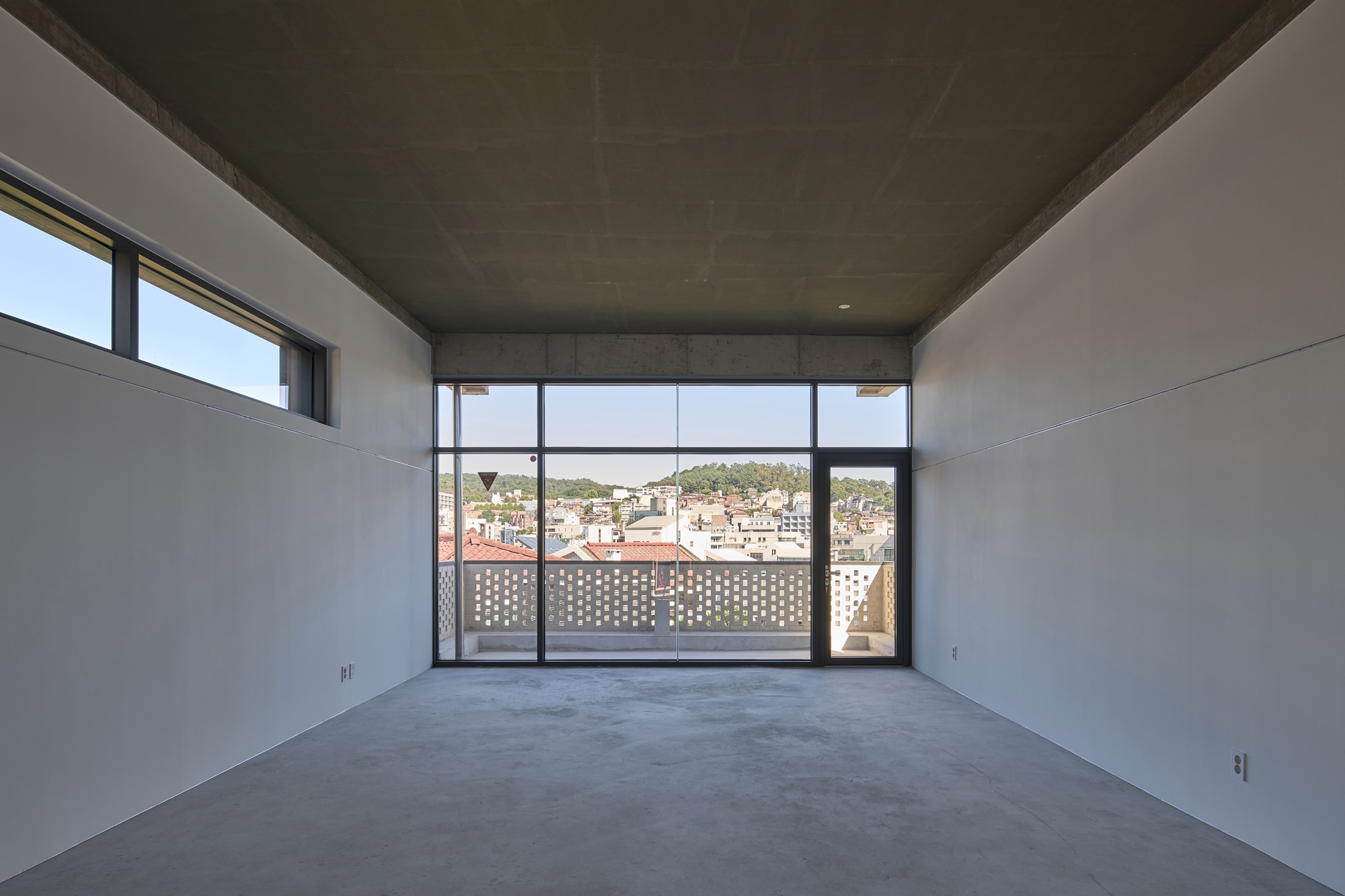Empty interior room with concrete ceiling and glazed wall opening to perforated screen and rooftops beyond