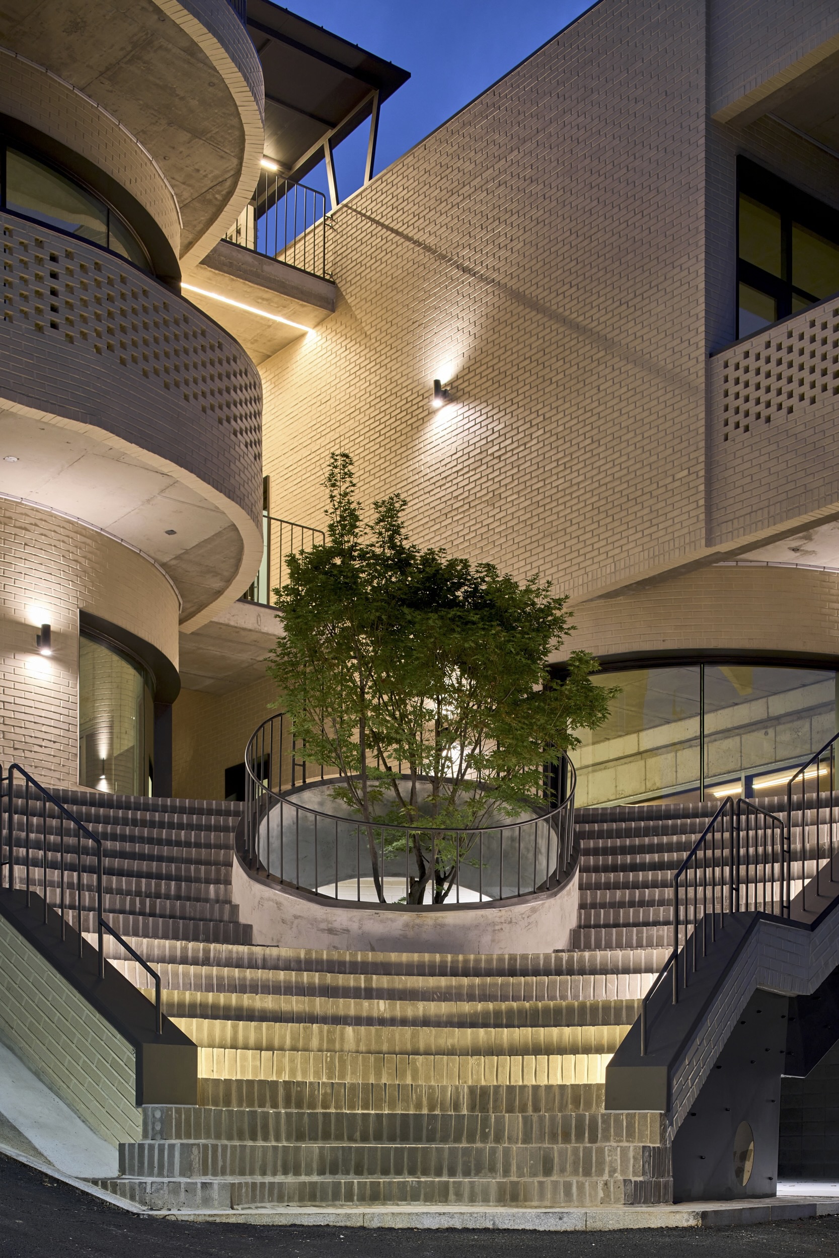 Curved exterior staircase descending to a courtyard with a central tree at twilight