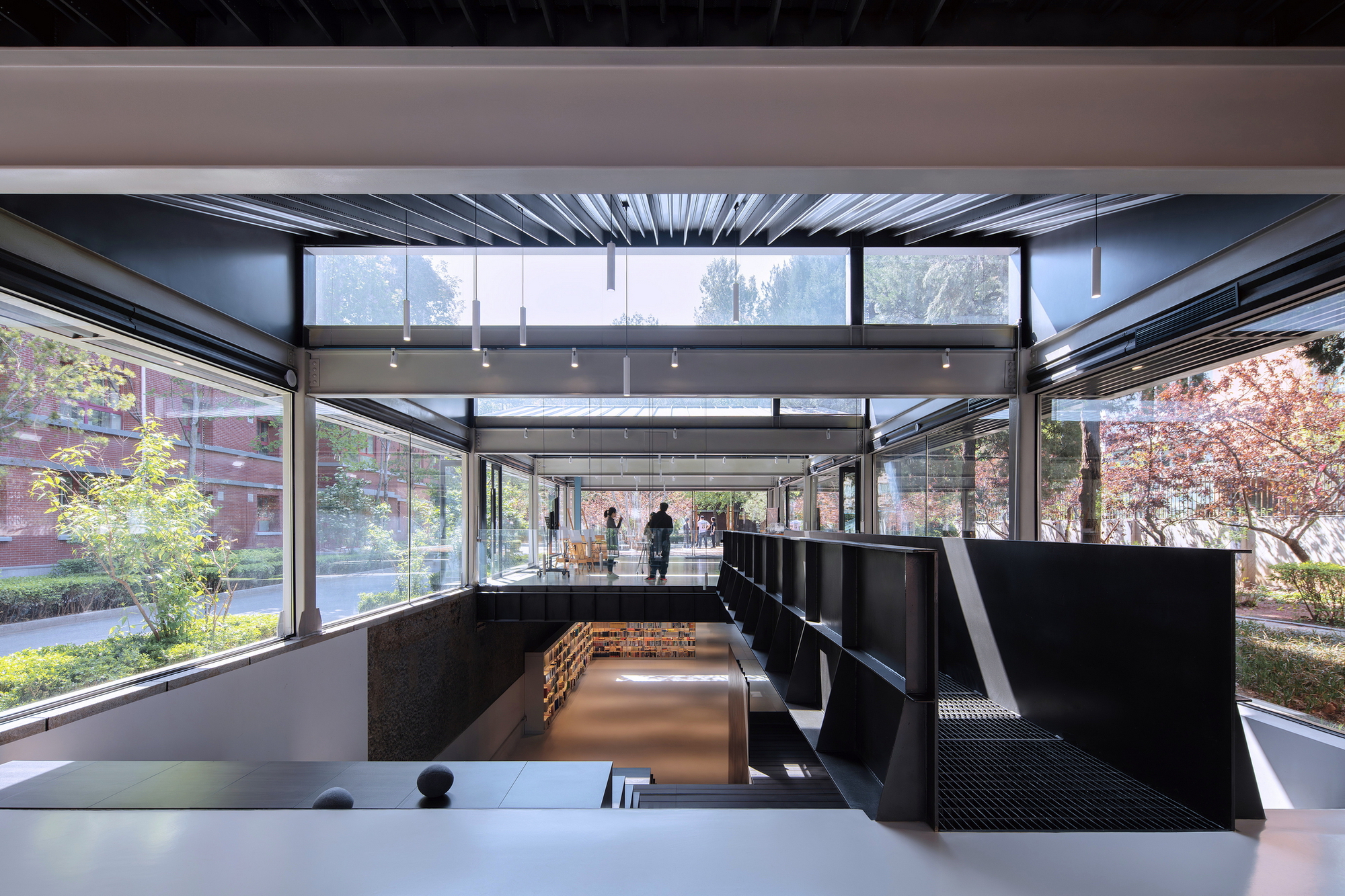 Split-level space with black shelving and perforated metal stairs framed by floor-to-ceiling glass overlooking courtyard gardens