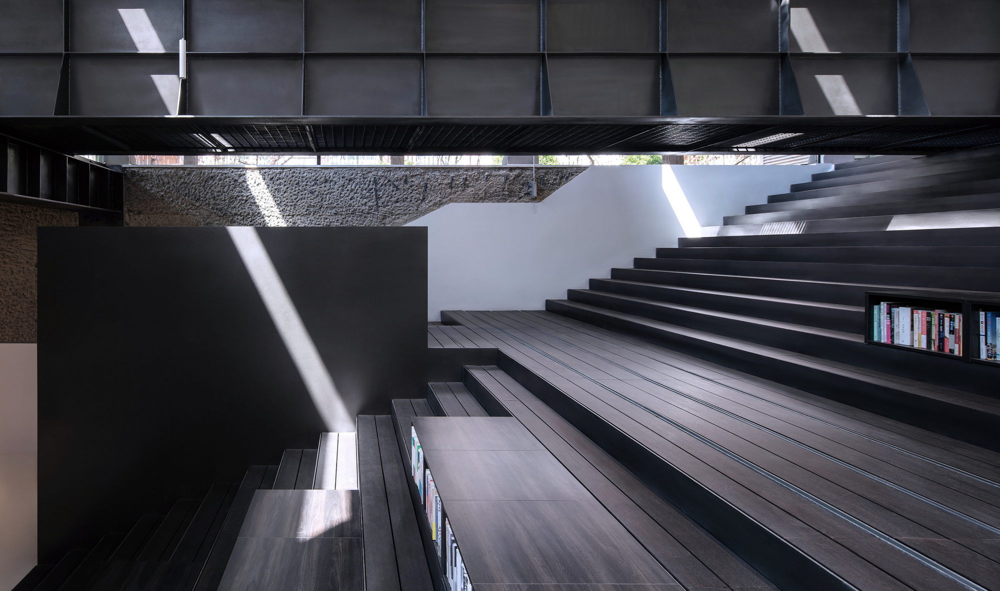 Looking up through the central atrium at black timber stair treads bridging between stone walls with sunlight streaming down