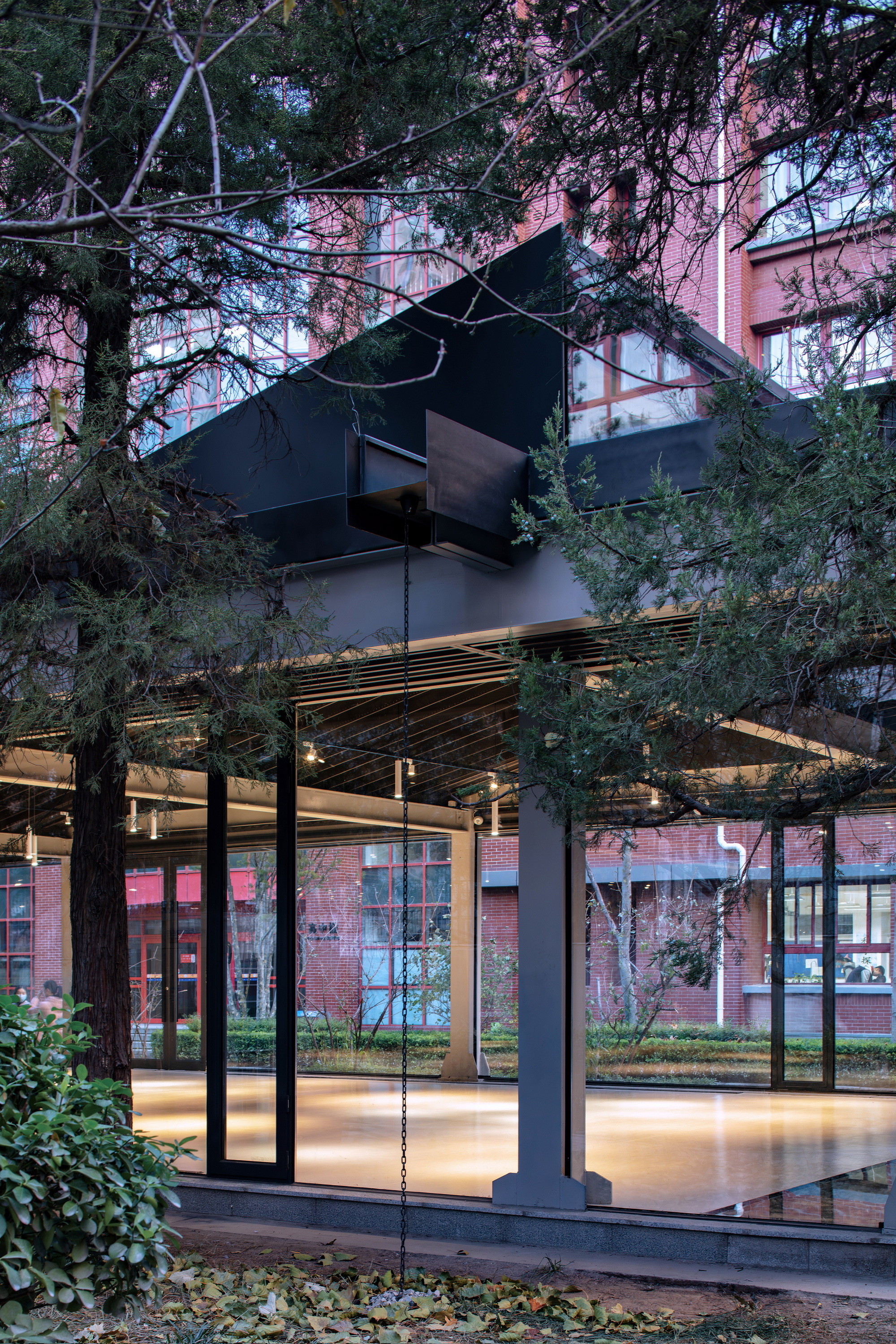 View through tree branches showing the illuminated timber ceiling and dark folded metal canopy above