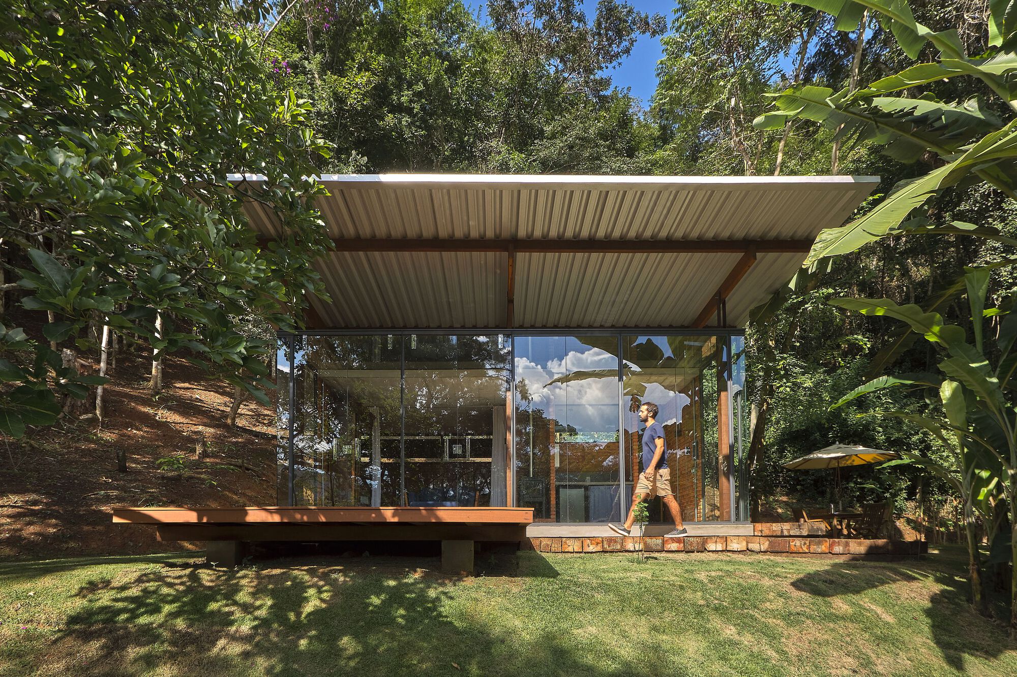 Glass pavilion with corrugated metal roof and timber deck set in a sloped clearing surrounded by trees