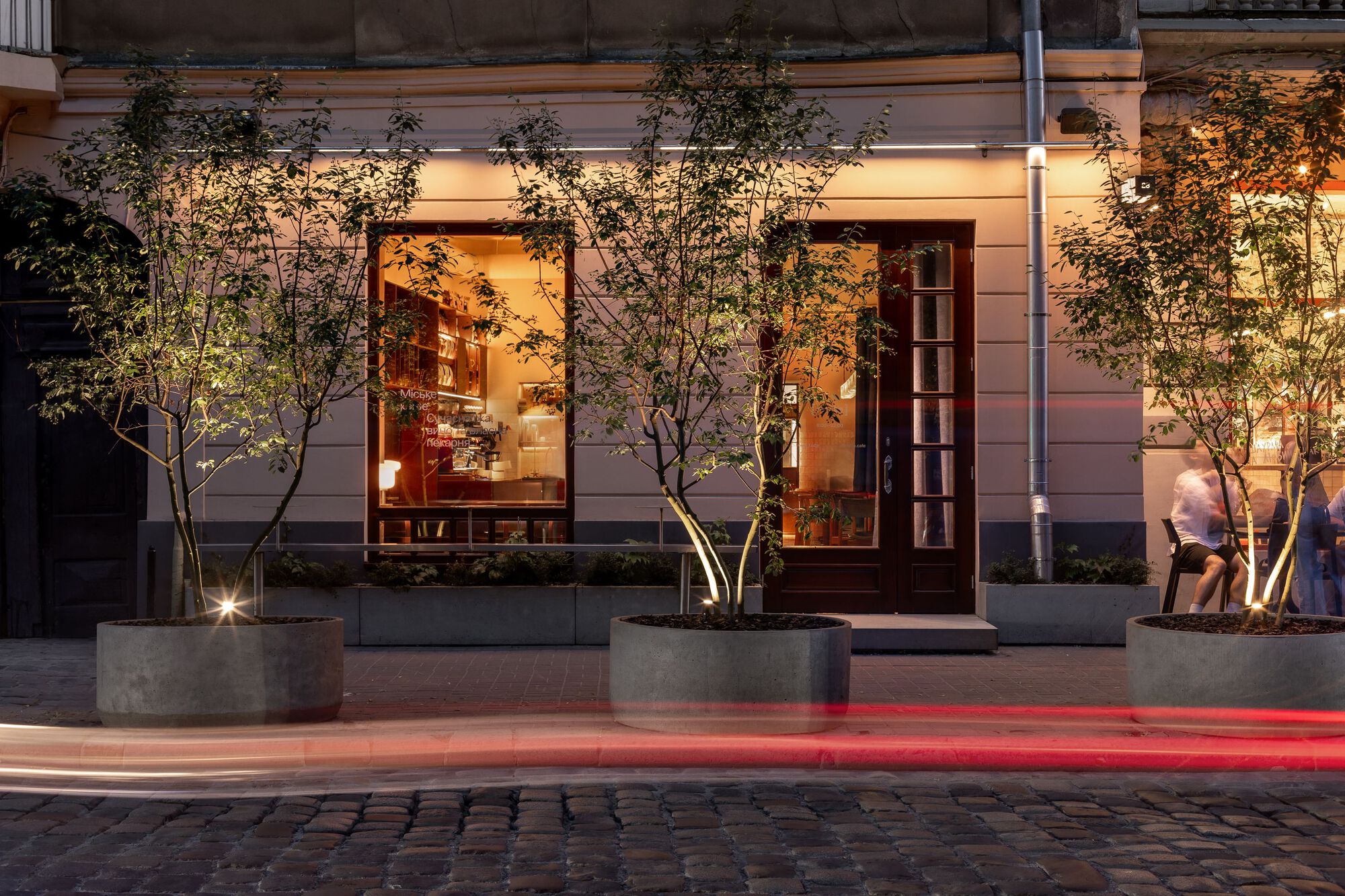 Street facade at dusk with concrete planters, uplighting on young trees and blurred pedestrians passing