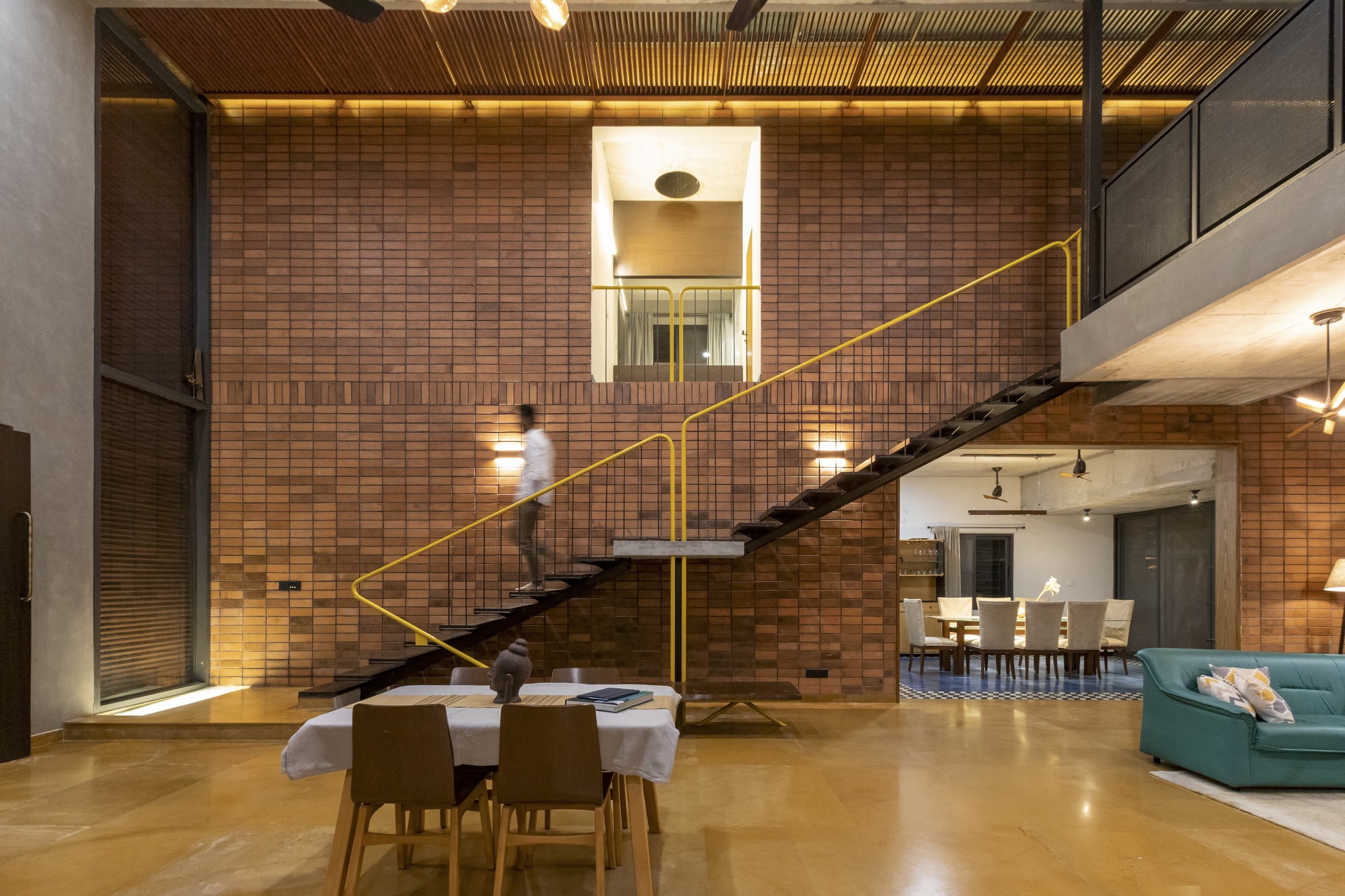 Double-height interior with floating staircase and yellow railing against a patterned brick wall at night