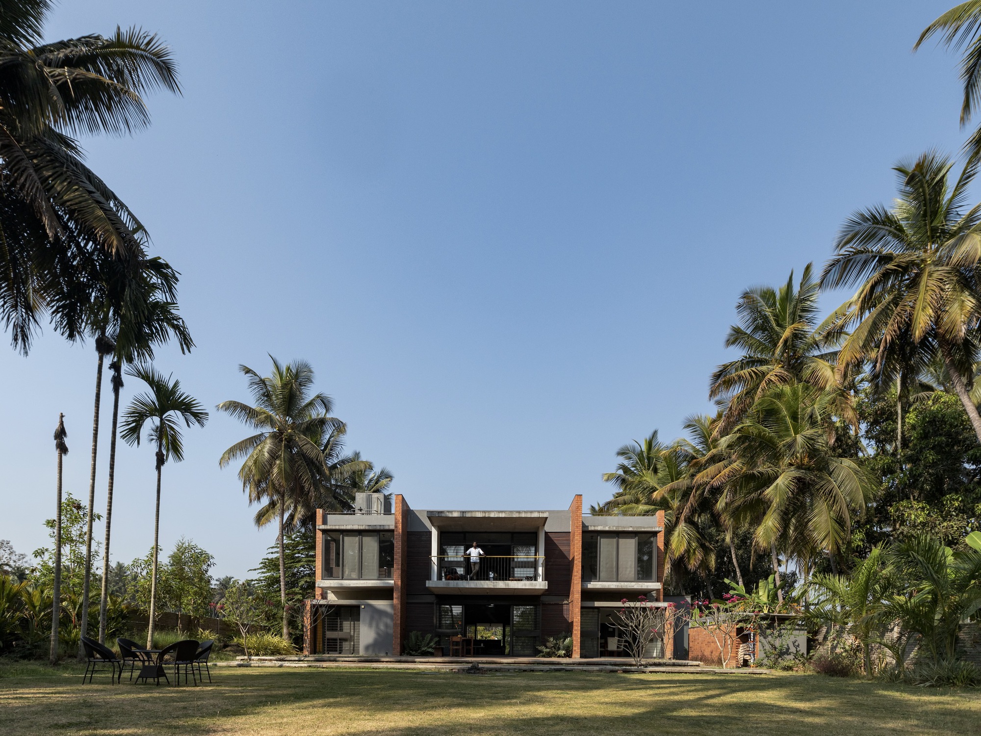 Garden elevation showing the two-story residence with terracotta volumes, glass walls, and palm trees against blue sky