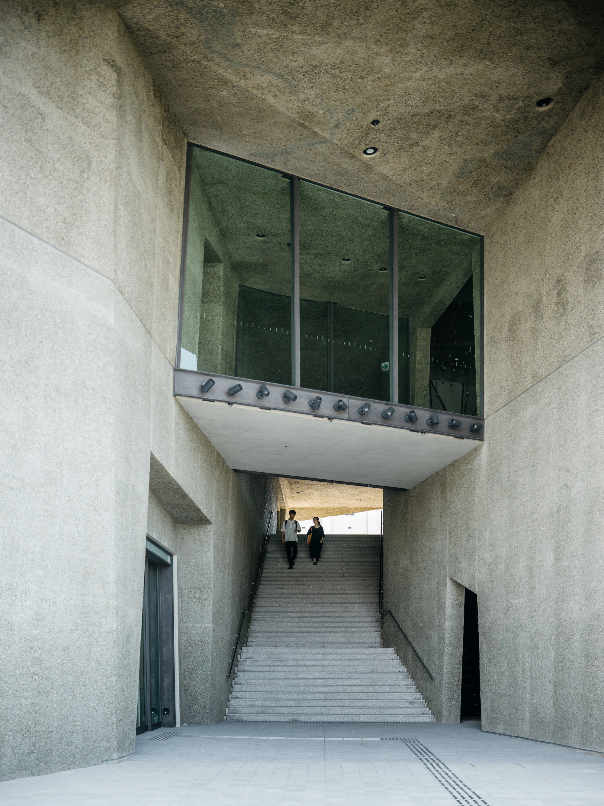 Concrete staircase ascending under a suspended glass box with two visitors climbing the steps