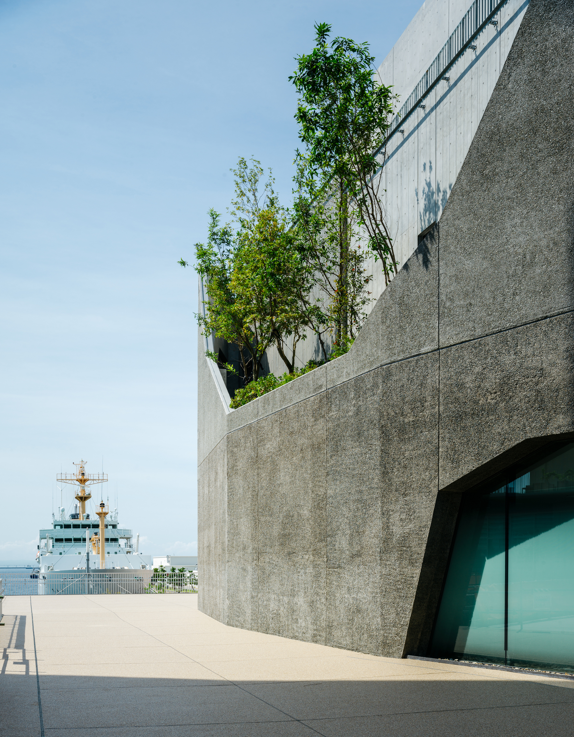 Concrete terrace with planted trees overlooking a harbor with a naval vessel in the distance