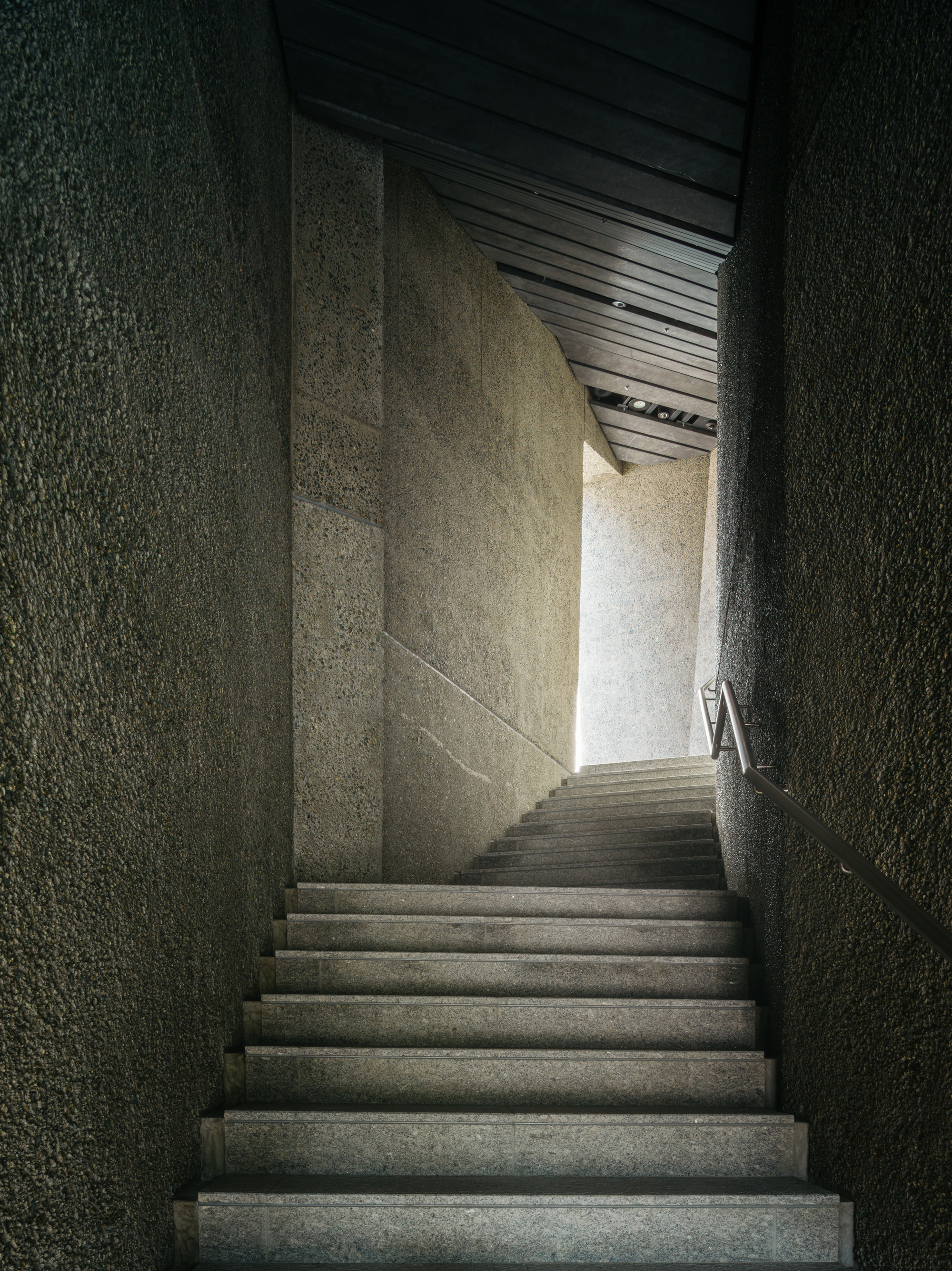 Narrow concrete staircase ascending between textured aggregate walls toward a bright opening above