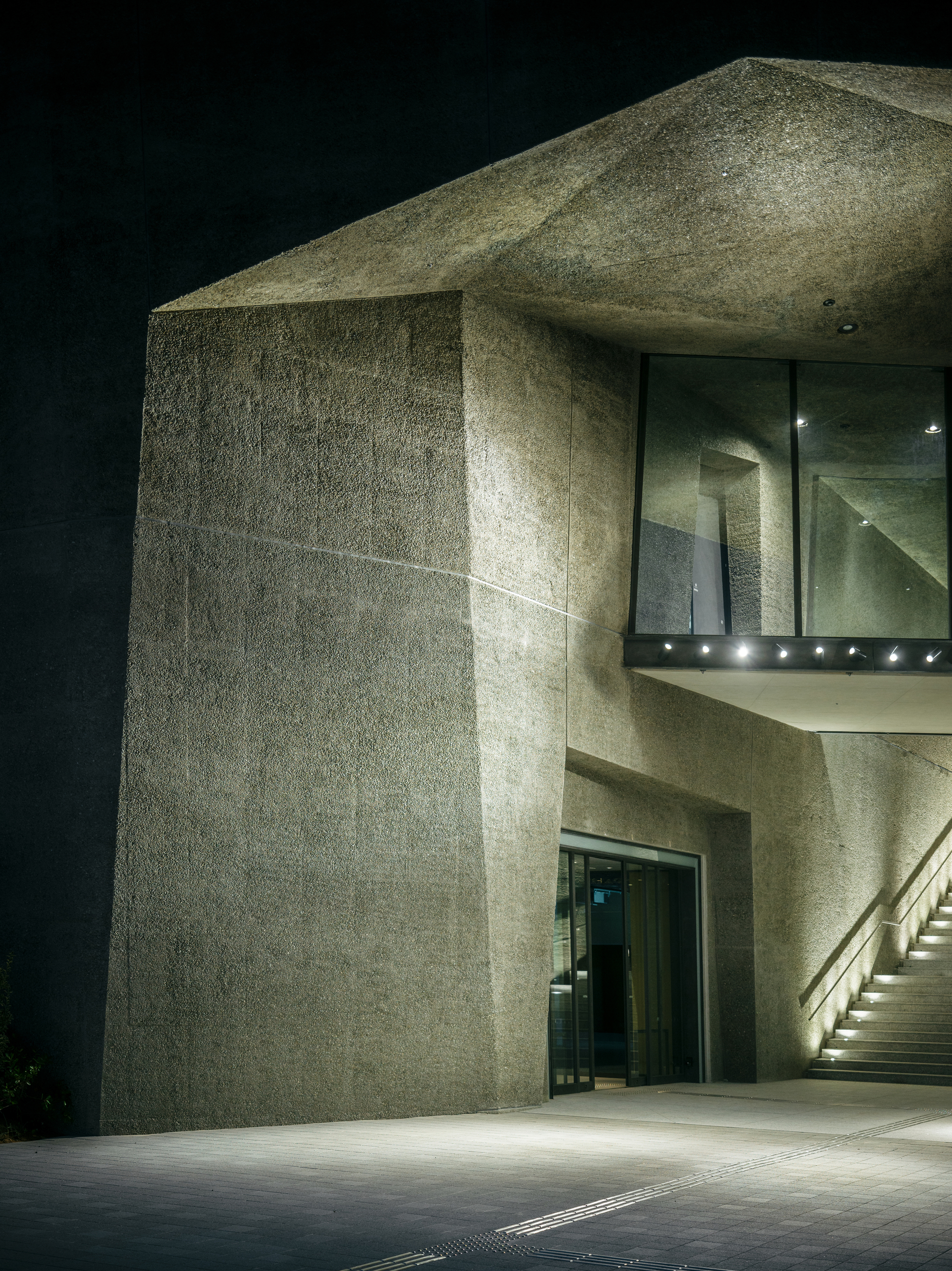 Textured concrete entrance canopy with glass walls and recessed lighting at night