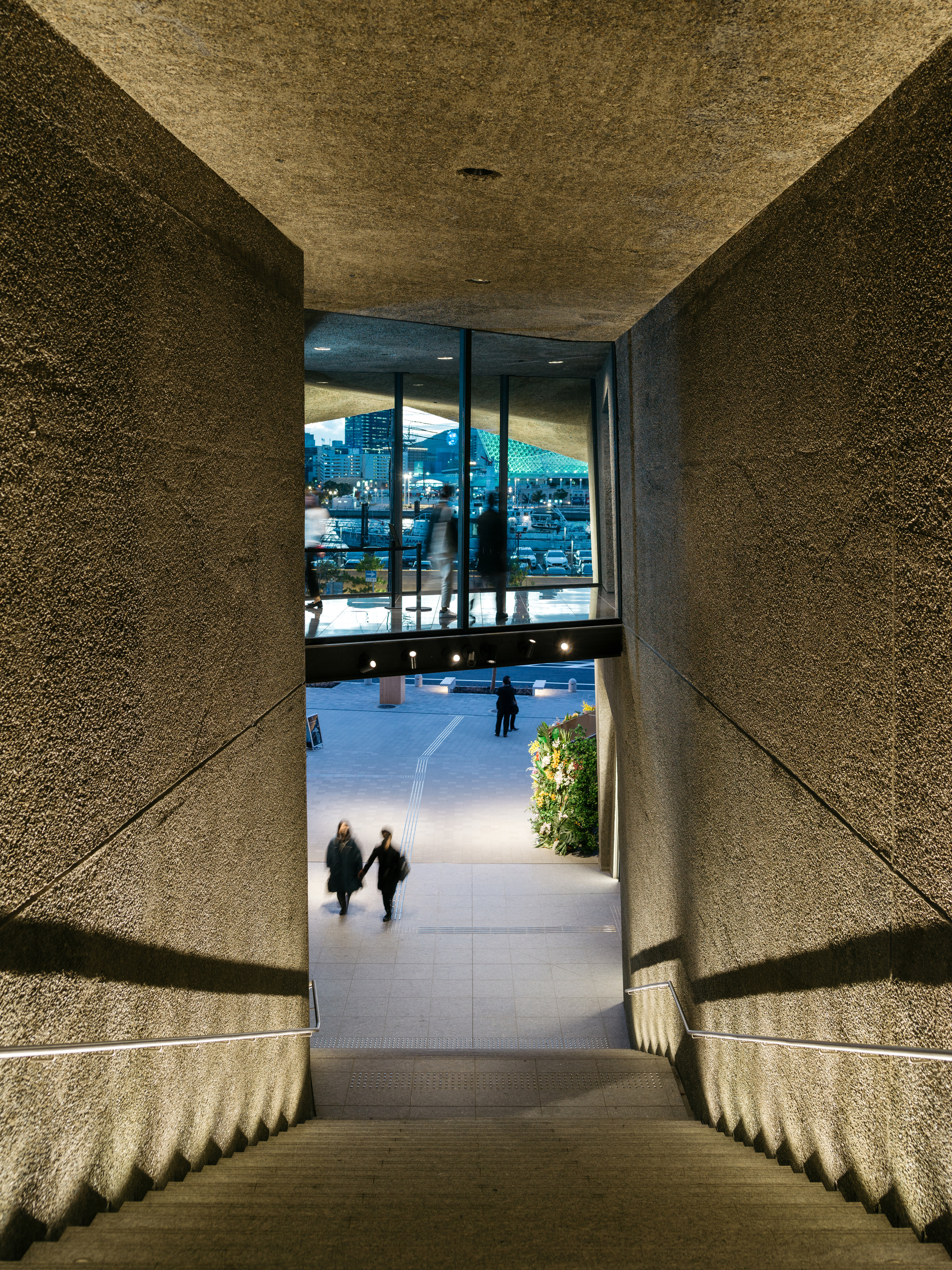 View down the stepped concrete passage with textured walls framing visitors walking toward the plaza at dusk