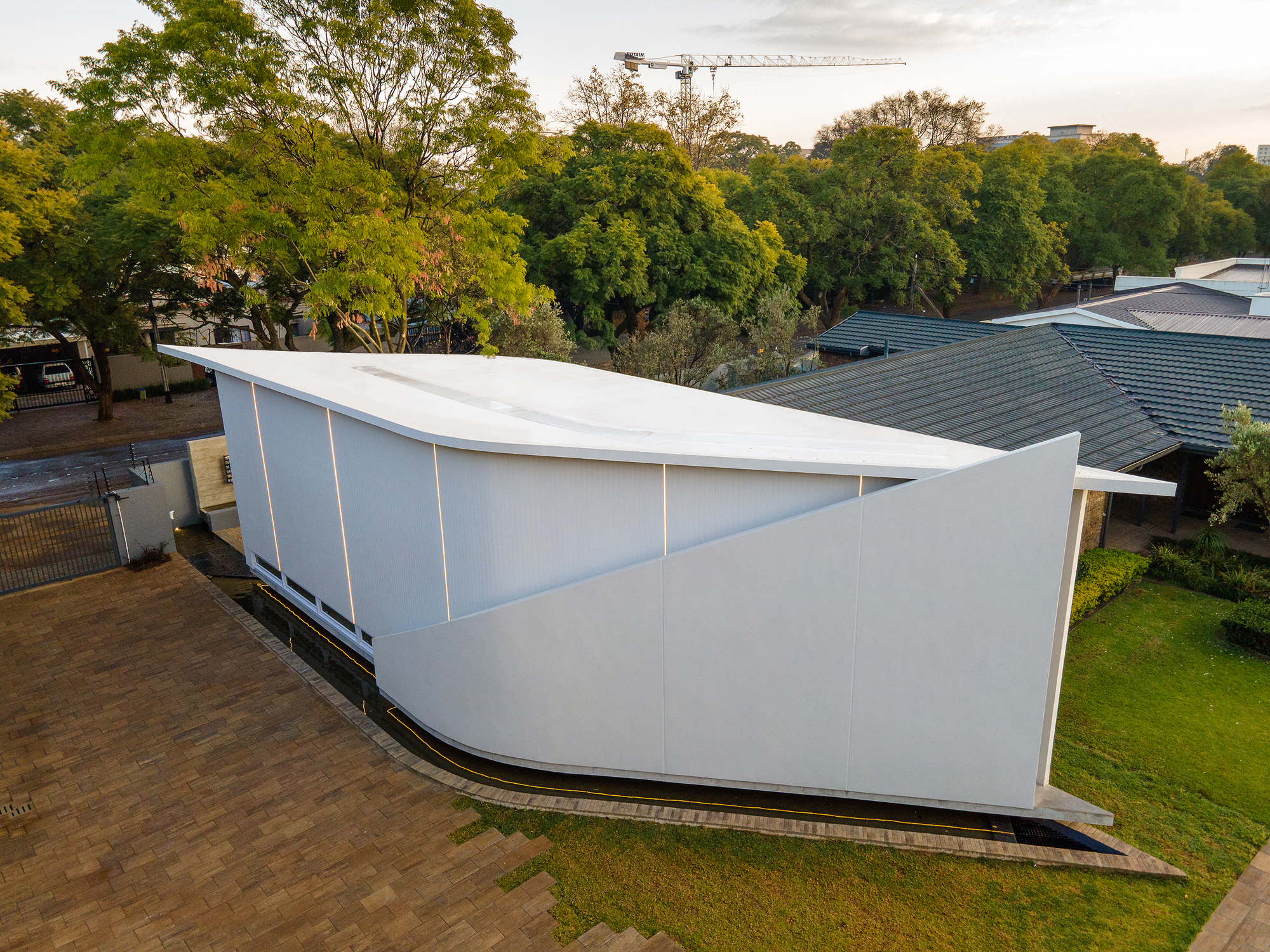Aerial view of the white curved volume with sloping roof set among mature trees at dusk