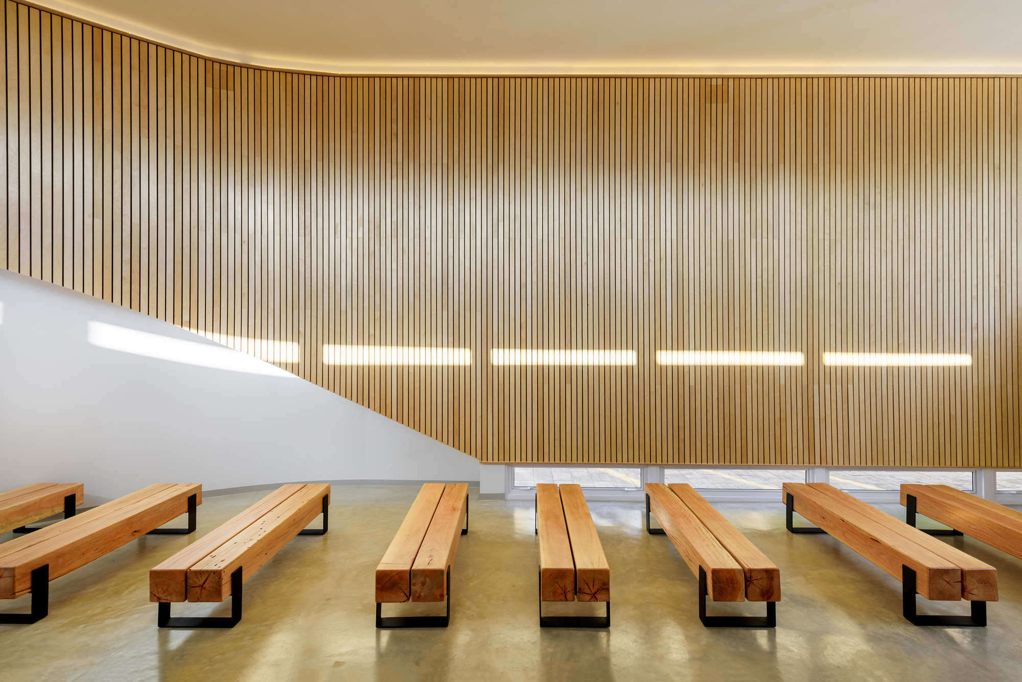 Vertical timber slat wall with five horizontal light strips above rows of wooden pews on concrete floor