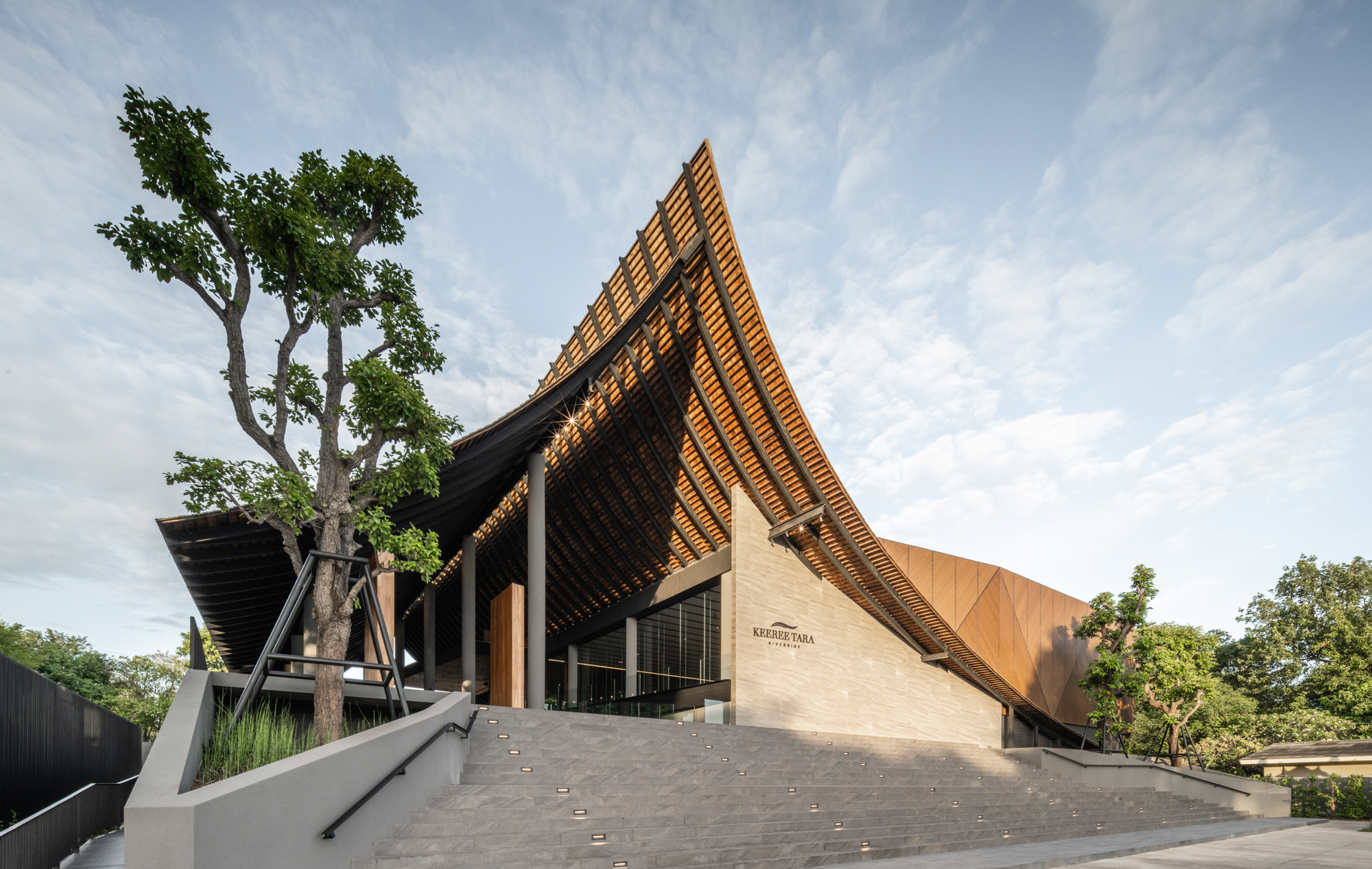 Approaching view of the sharply pitched timber roof and limestone facade beside a mature tree