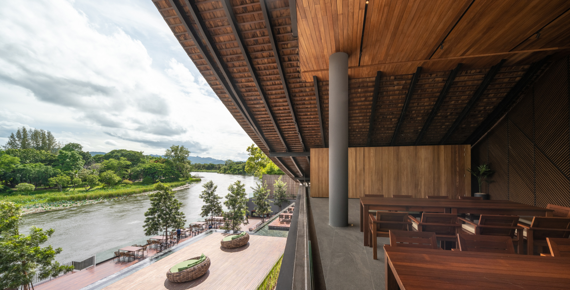 Cantilevered roof with exposed timber structure and cylindrical column overlooking a riverbank terrace and distant mountains