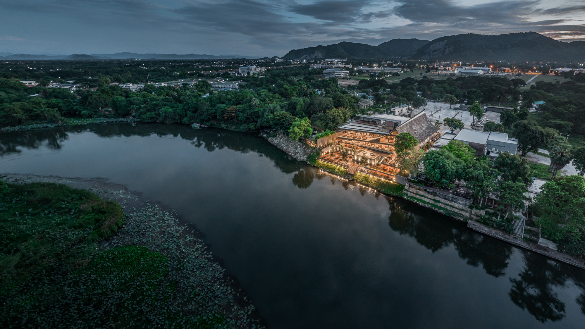 Drone view of illuminated timber pavilion on river's edge at dusk with mountains in background