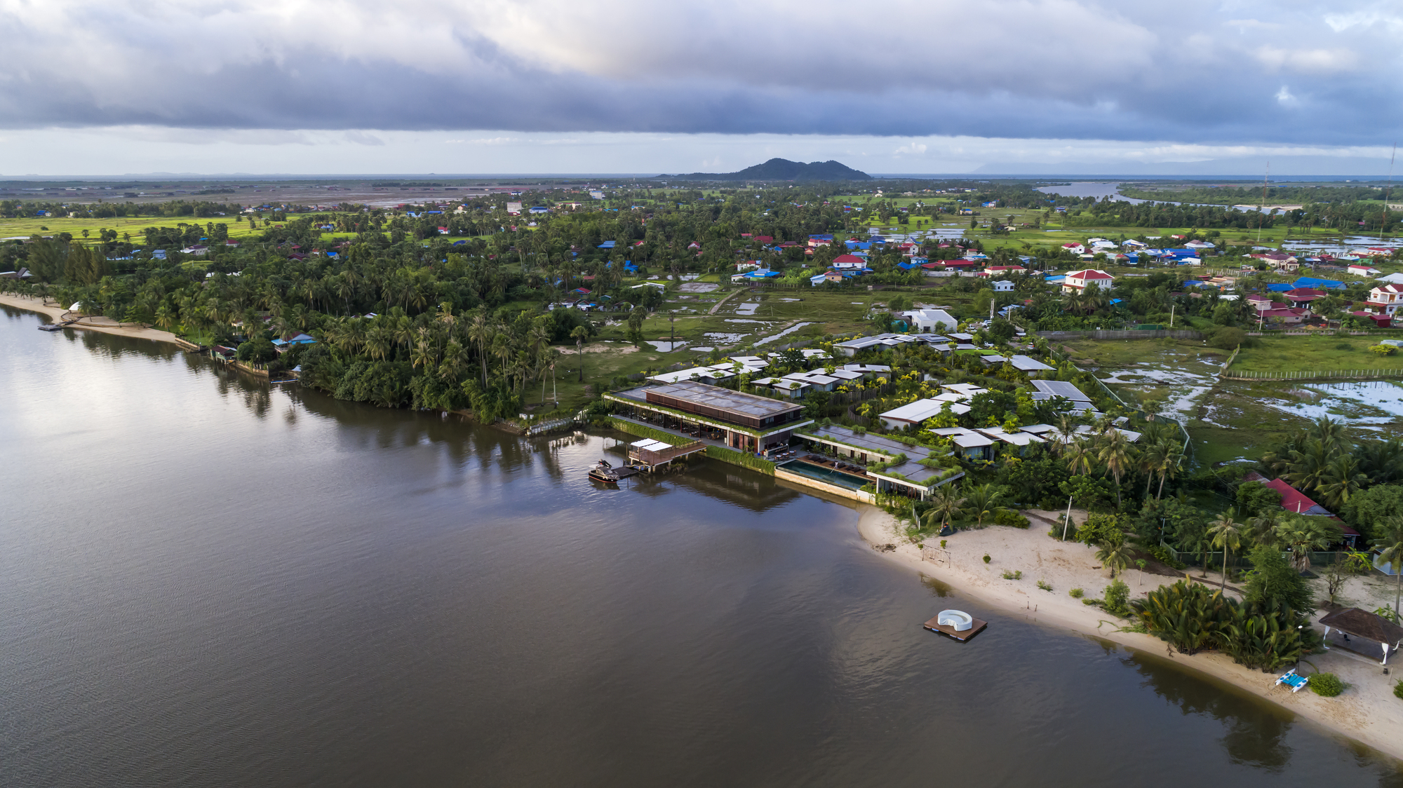 Aerial view of the horizontal volumes along the riverbank with palm trees and distant mountains under storm clouds