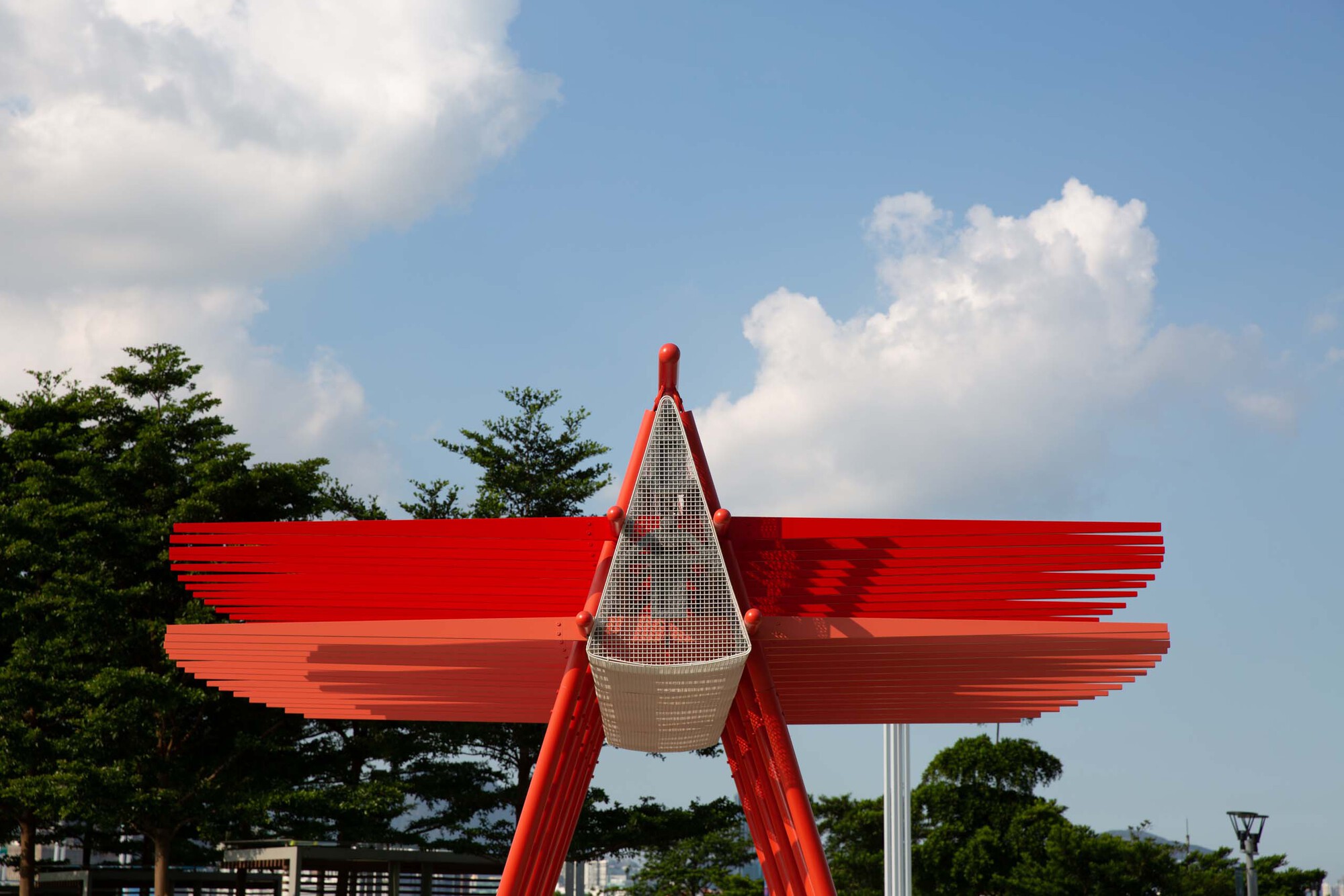 Close-up of the red steel canopy with mesh enclosure and layered horizontal fins against a partly cloudy sky