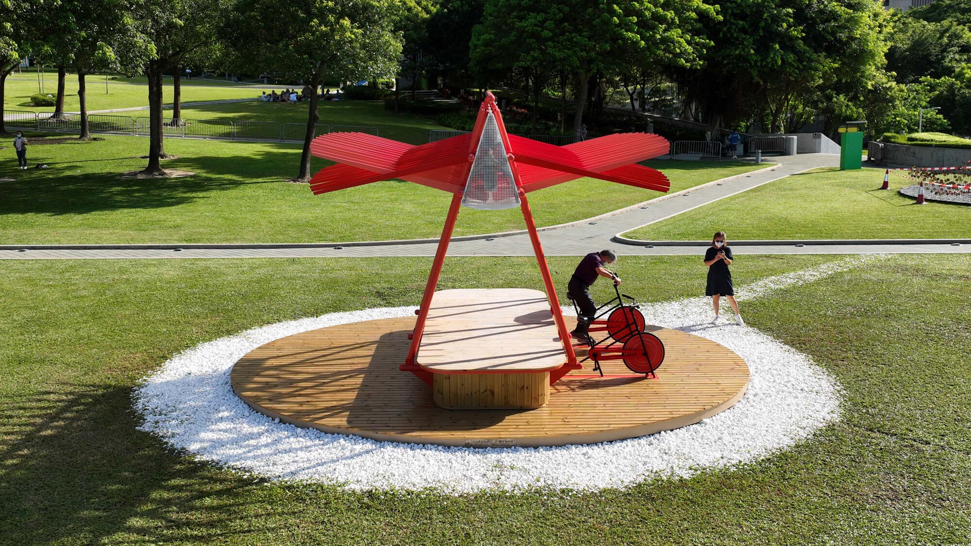 Red steel pavilion with timber deck and white gravel ring in a sunlit park lawn