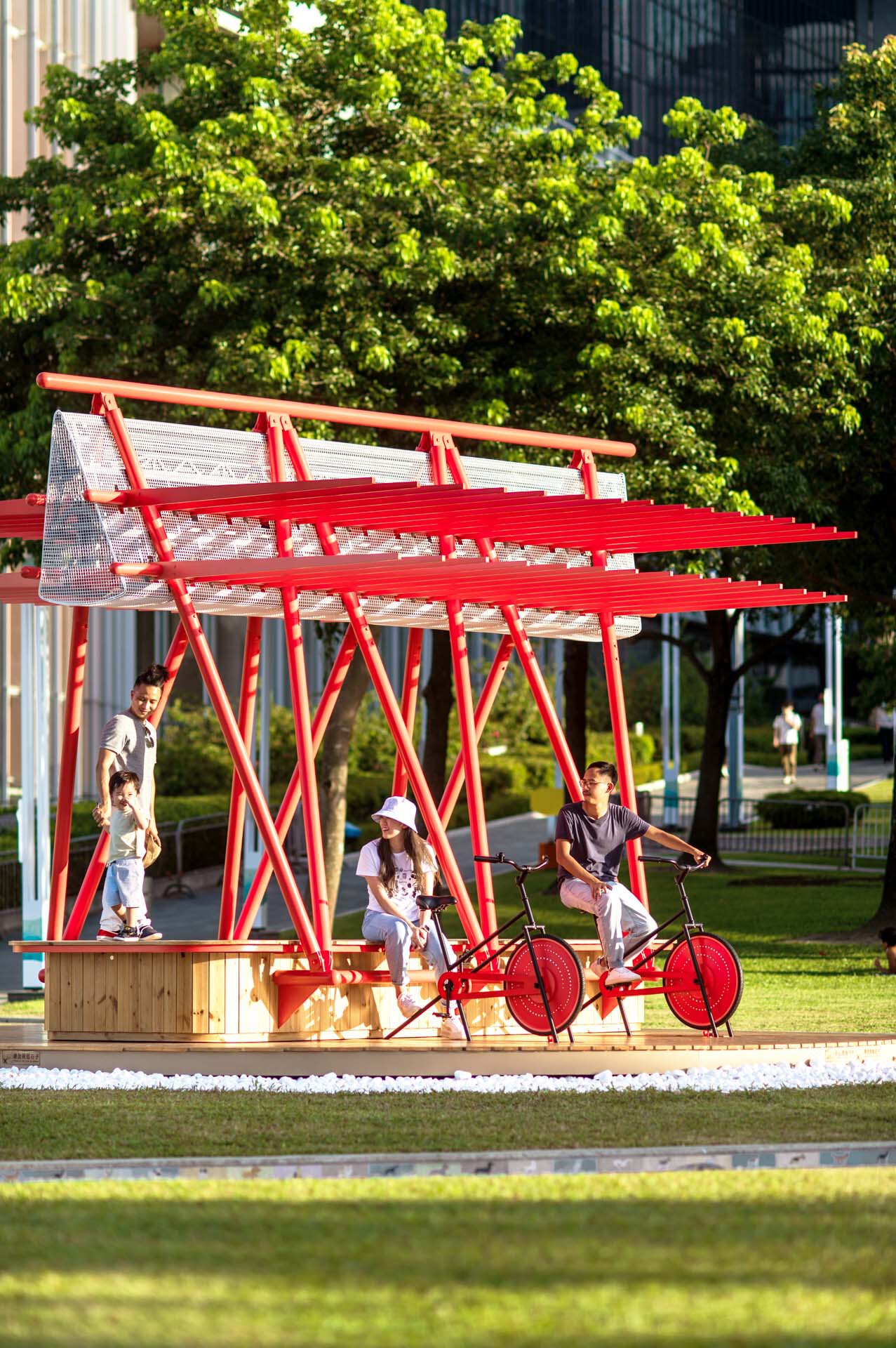 Red steel canopy sheltering a wooden seating platform with visitors on a bicycle installation