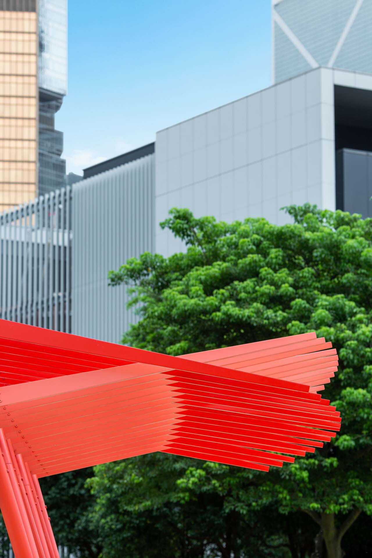 Close-up of layered red steel slat canopy against green trees and glass tower facades