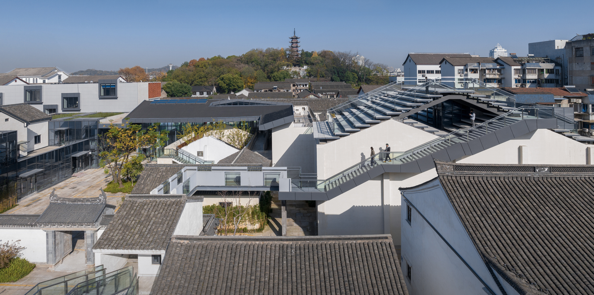 Aerial view of interconnected courtyards with white volumes and grey-tiled roofs among surrounding neighborhood buildings