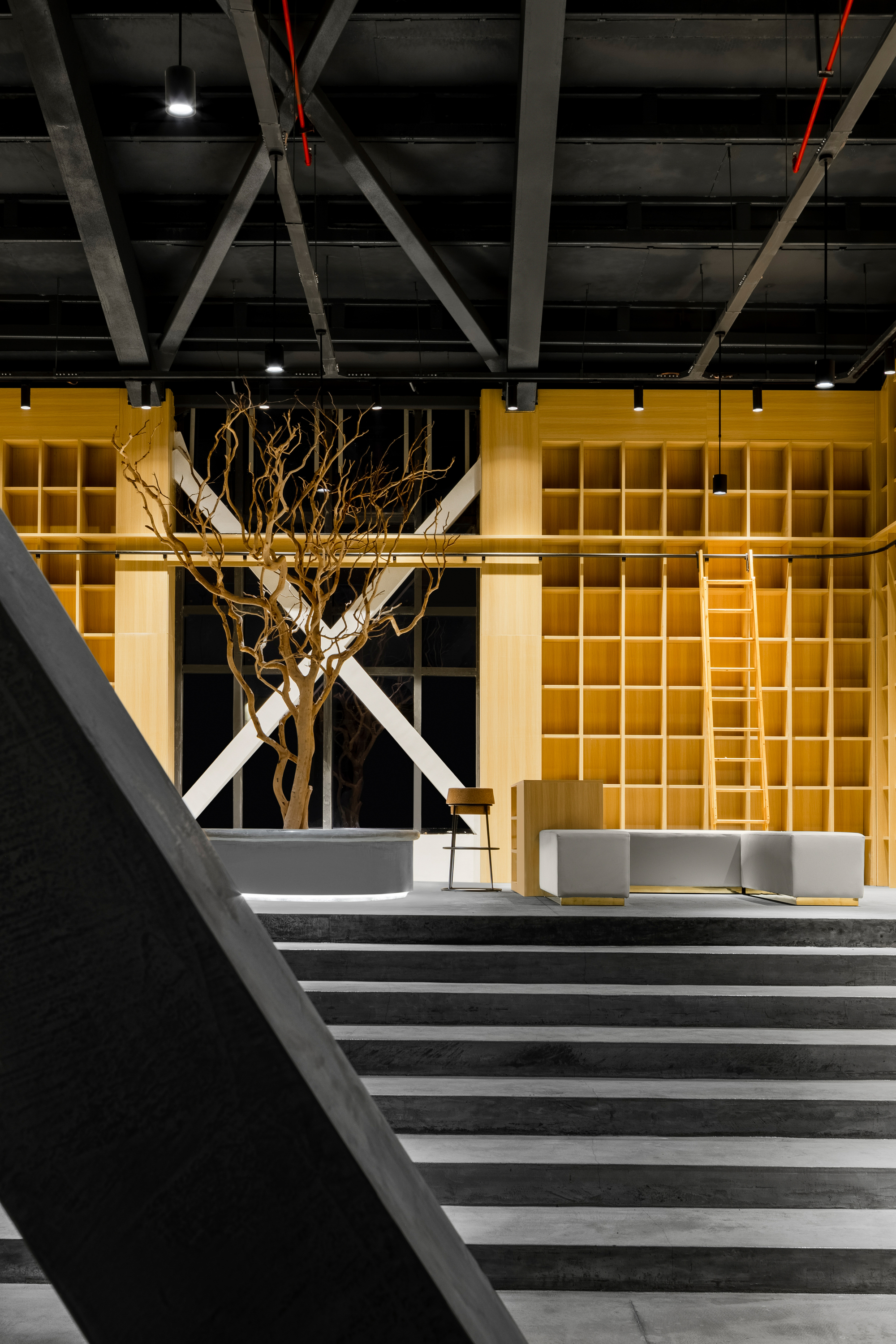 Interior lobby with exposed diagonal steel trusses above yellow illuminated shelving wall and sculptural tree branch