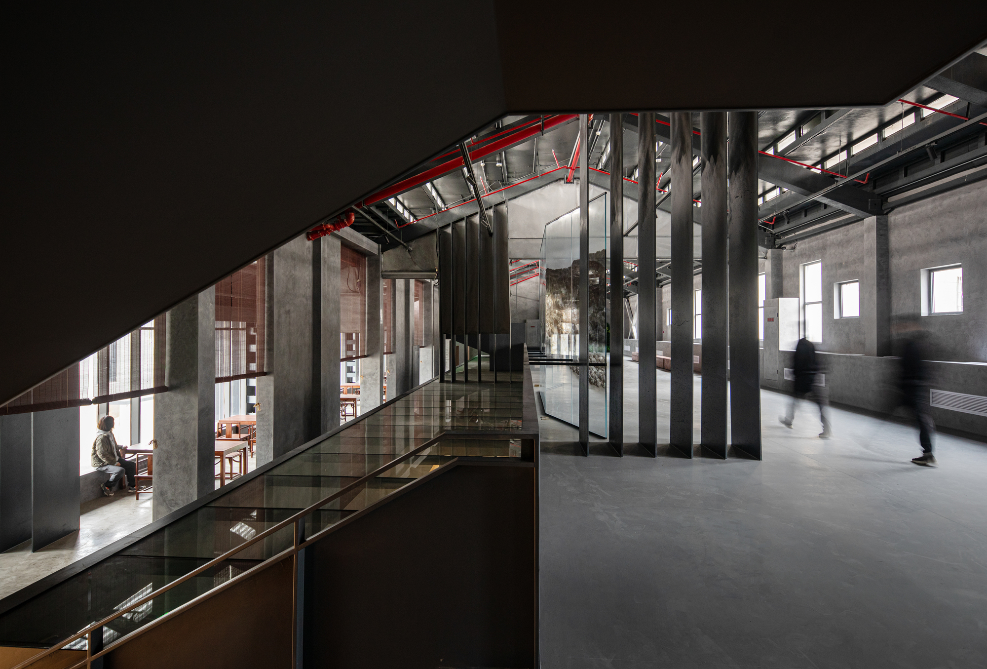 Mezzanine corridor with red steel trusses and glass balustrade overlooking exhibition spaces below