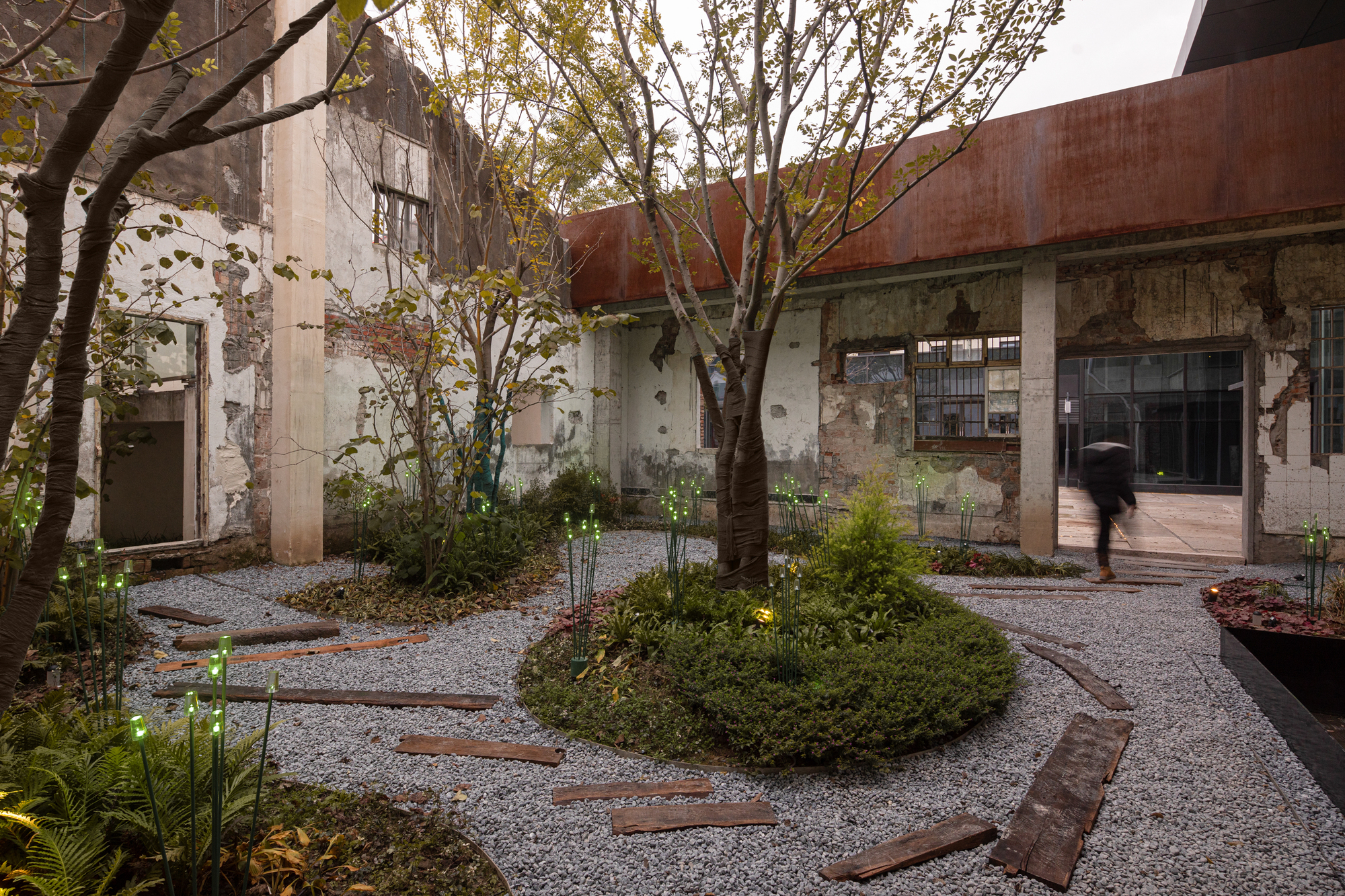 Gravel courtyard with circular planted beds and deciduous trees among weathered concrete walls