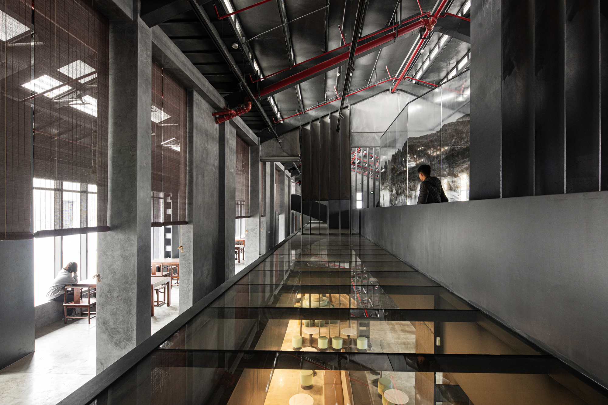 Glass-floored bridge suspended between concrete columns with red conduit overhead and a figure standing at the end
