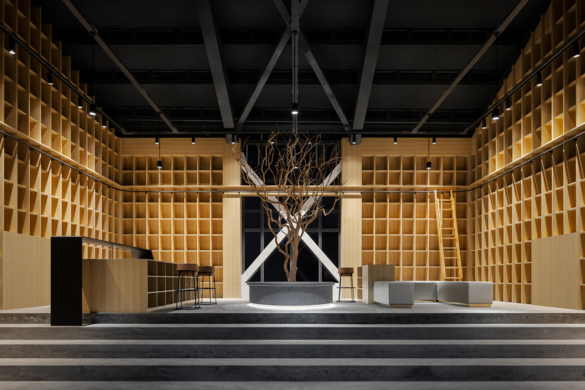 Gallery space with gridded timber wall shelving and a bare tree in a circular planter beneath exposed steel trusses