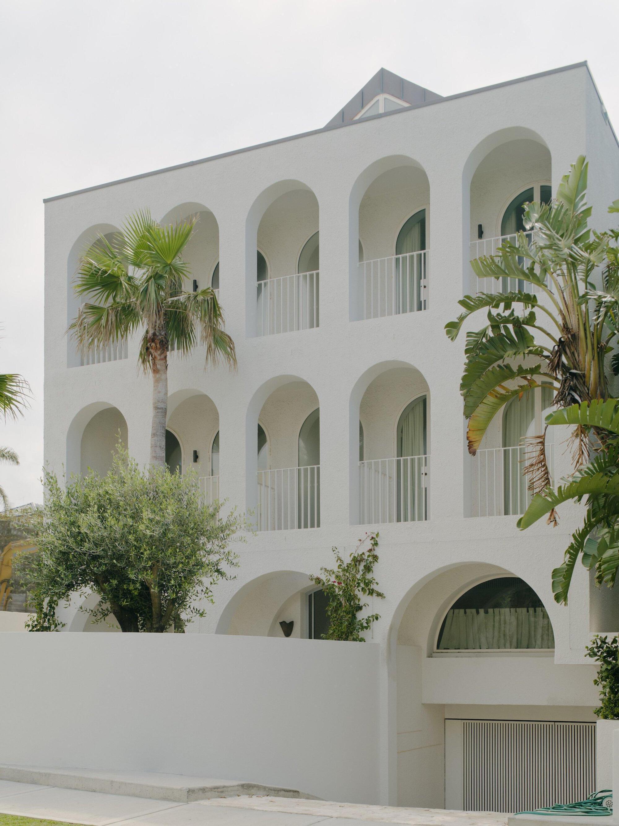 White stucco facade with arched openings and white balustrades, framed by palms and tropical foliage