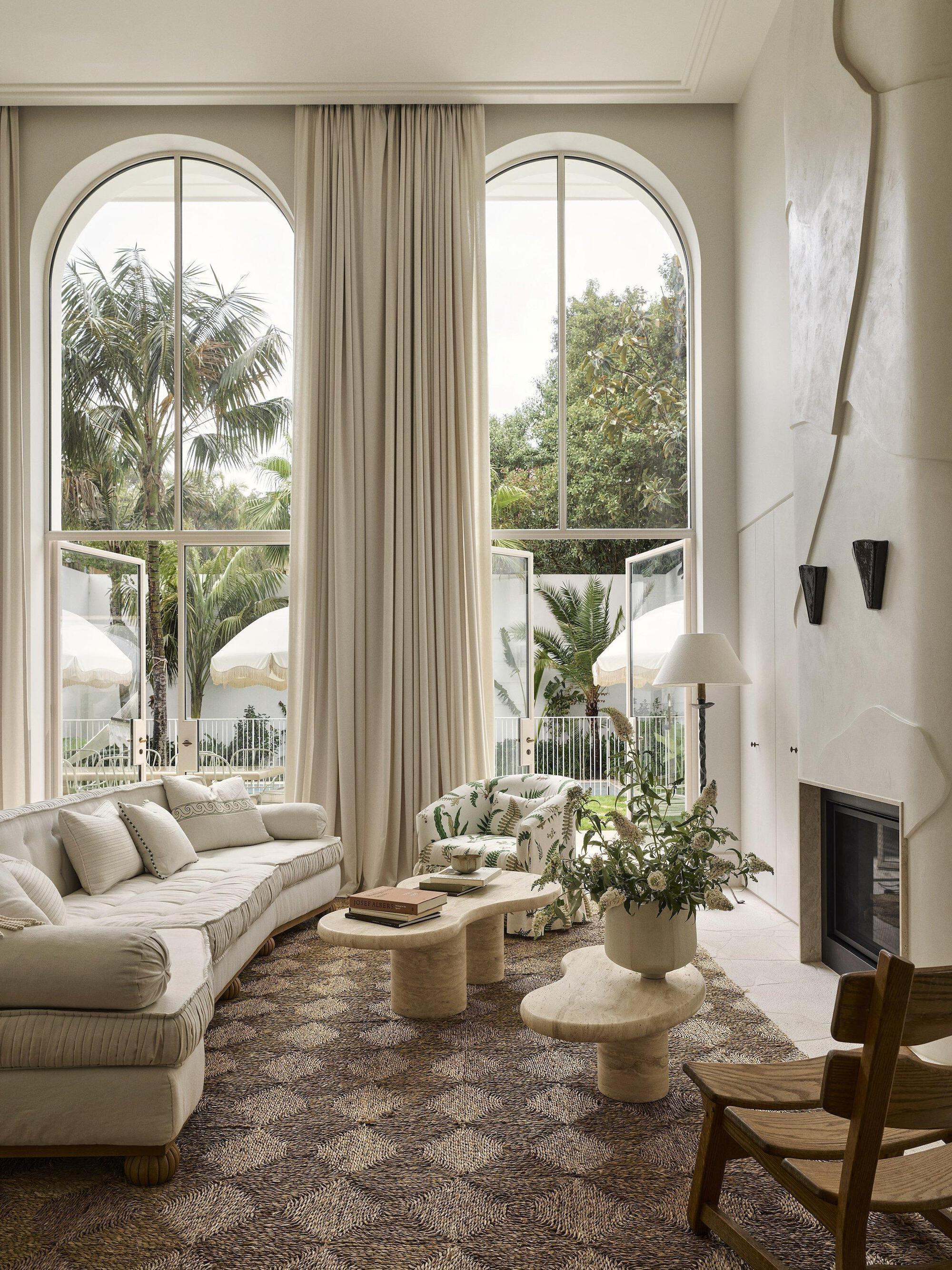 Double-height living room with arched windows framing palm trees and a curved plaster fireplace