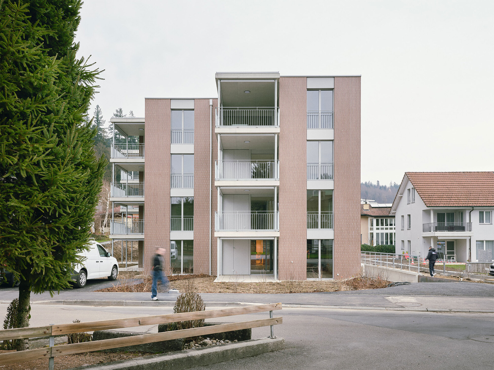 Street view of the four-storey apartment block with ribbed facade and white balconies as a figure walks past